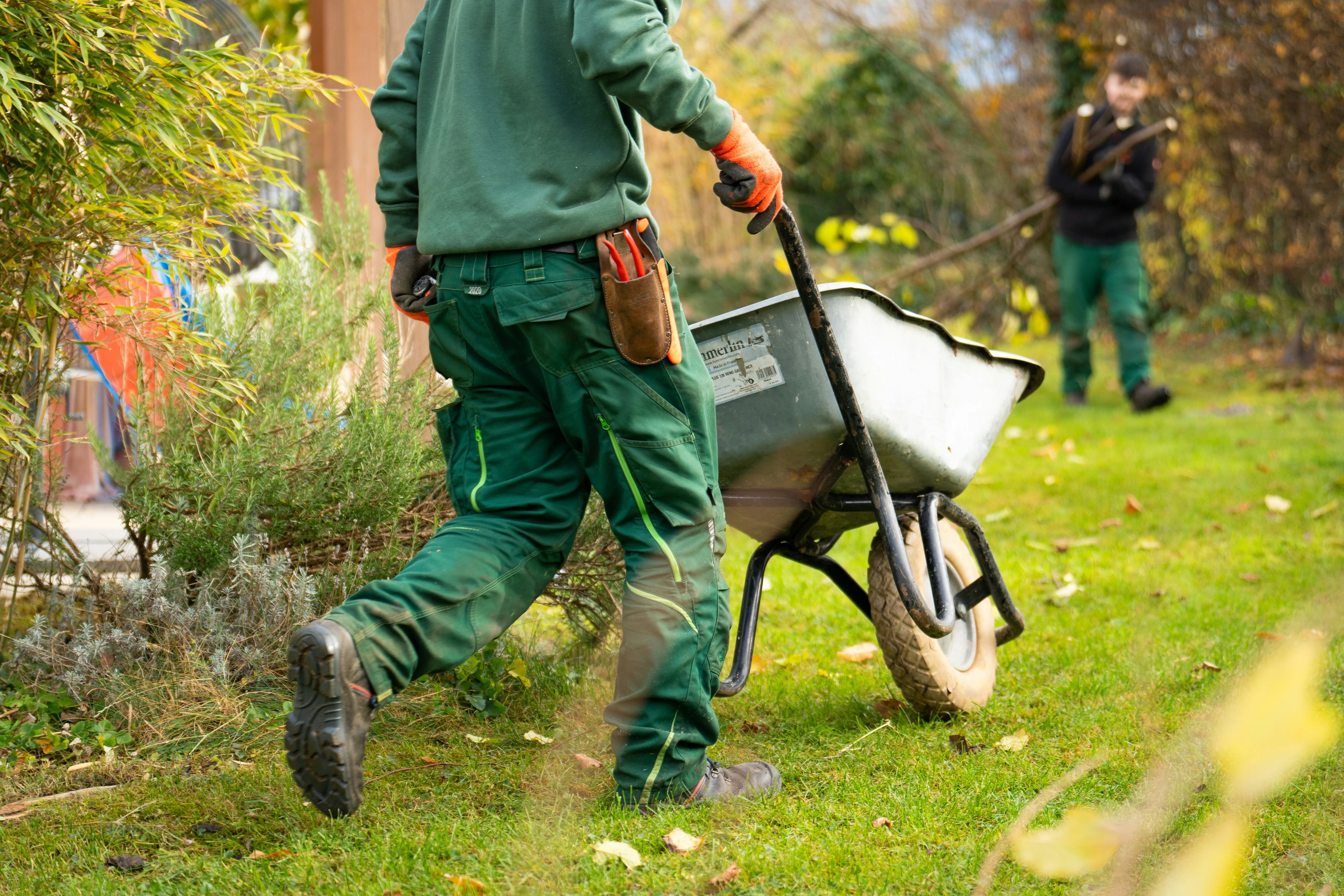 Two landscapers working in a garden, one with a wheelbarrow, showing groundworks or clearance.