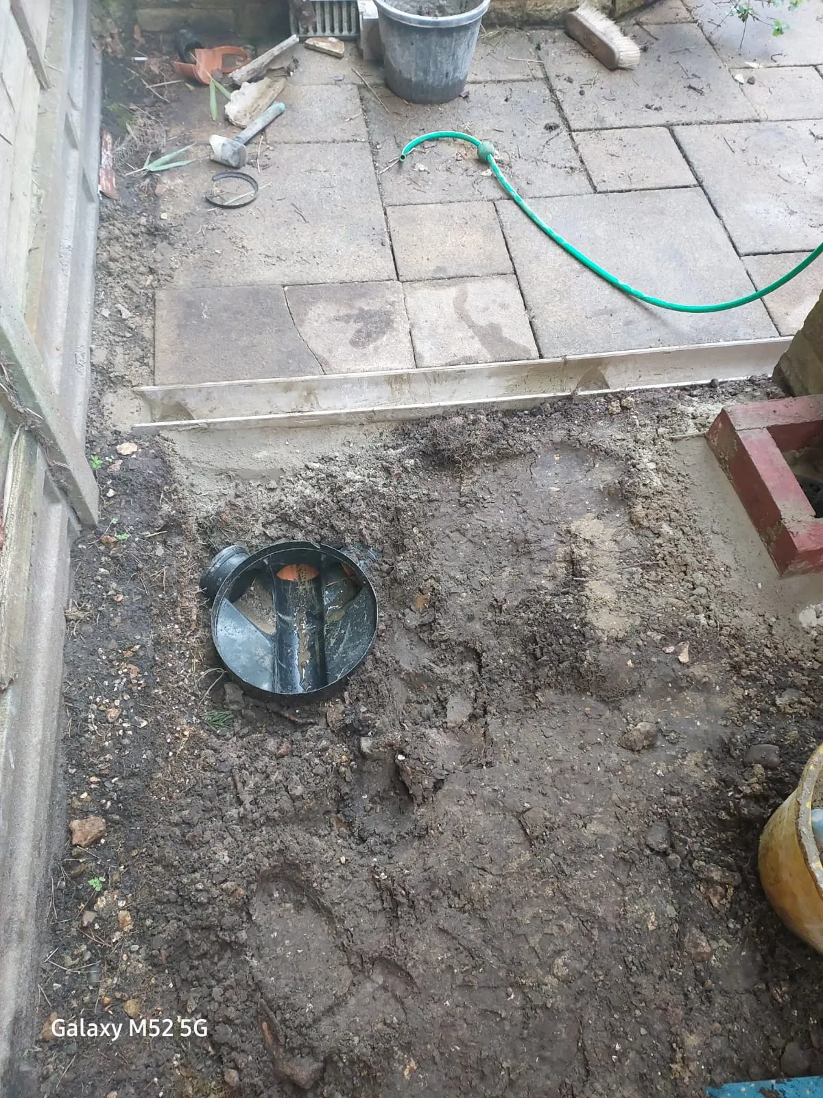 A black circular drainage component being installed in a muddy trench next to a paved patio, with a green hose nearby.