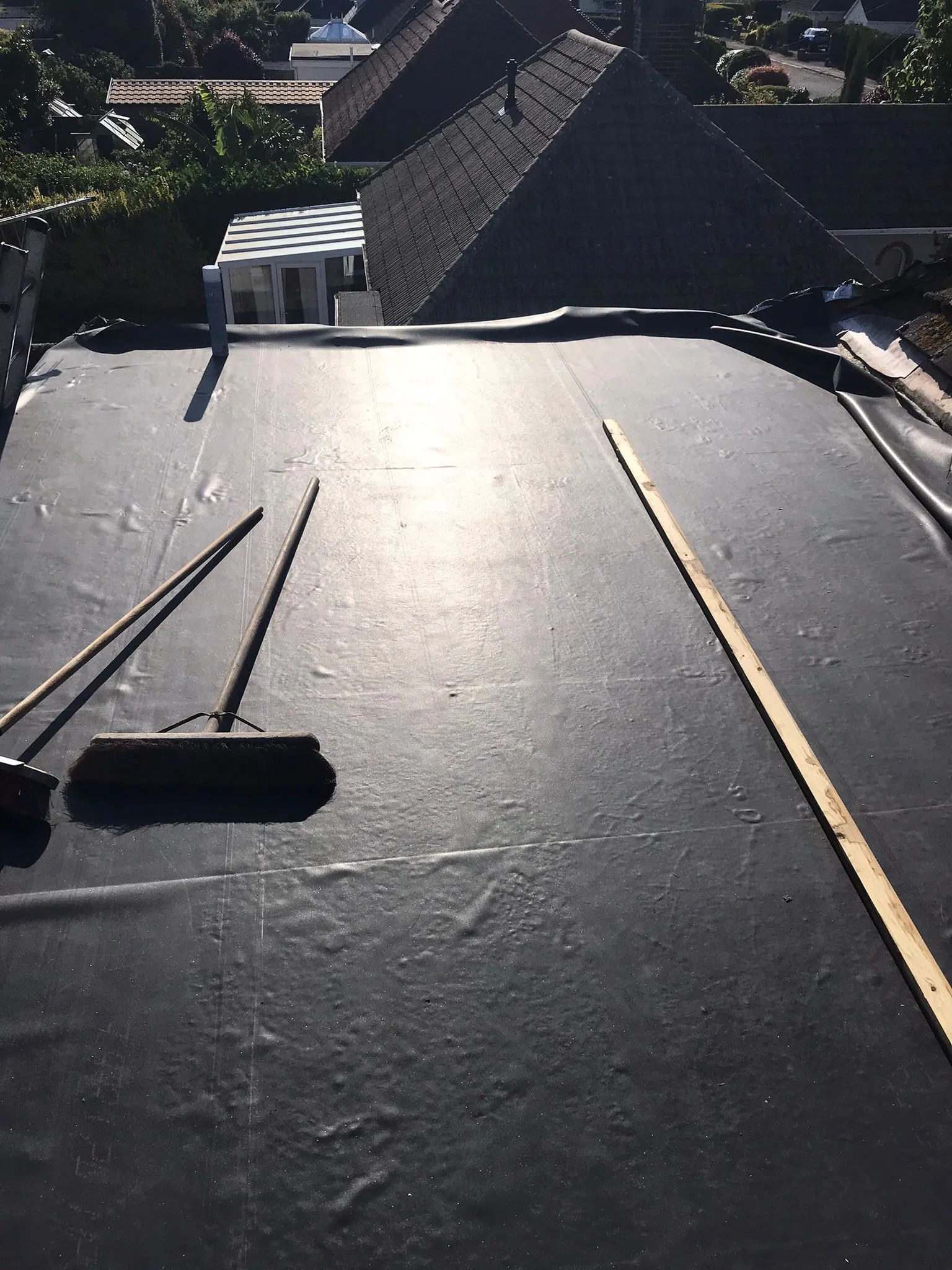A flat roof covered with a dark membrane, with brooms and a wooden batten, indicating active roofing work. Residential rooftops are visible in the distance.