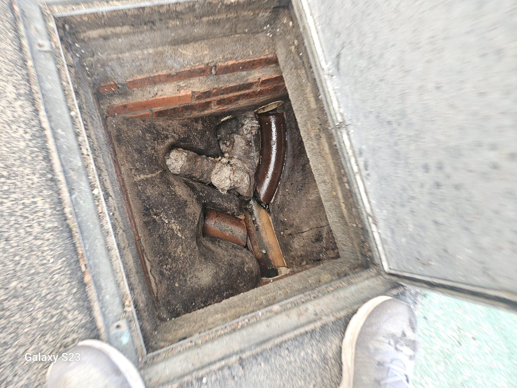 View into an open manhole showing a drain pipe heavily blocked with debris and waste, indicating a drainage problem.