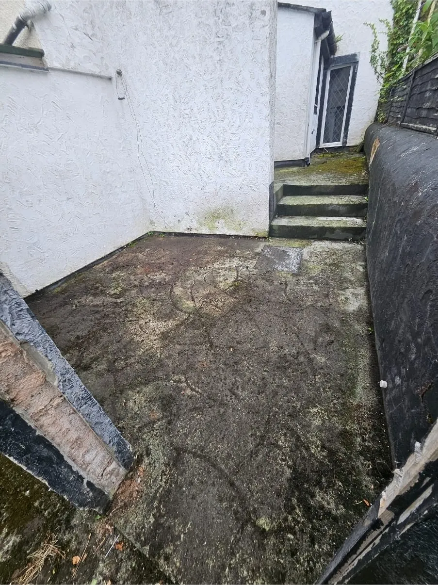 A dirty, neglected paved patio area with moss and grime on the ground and walls, showing an area ready for garden clearance or rubbish removal.