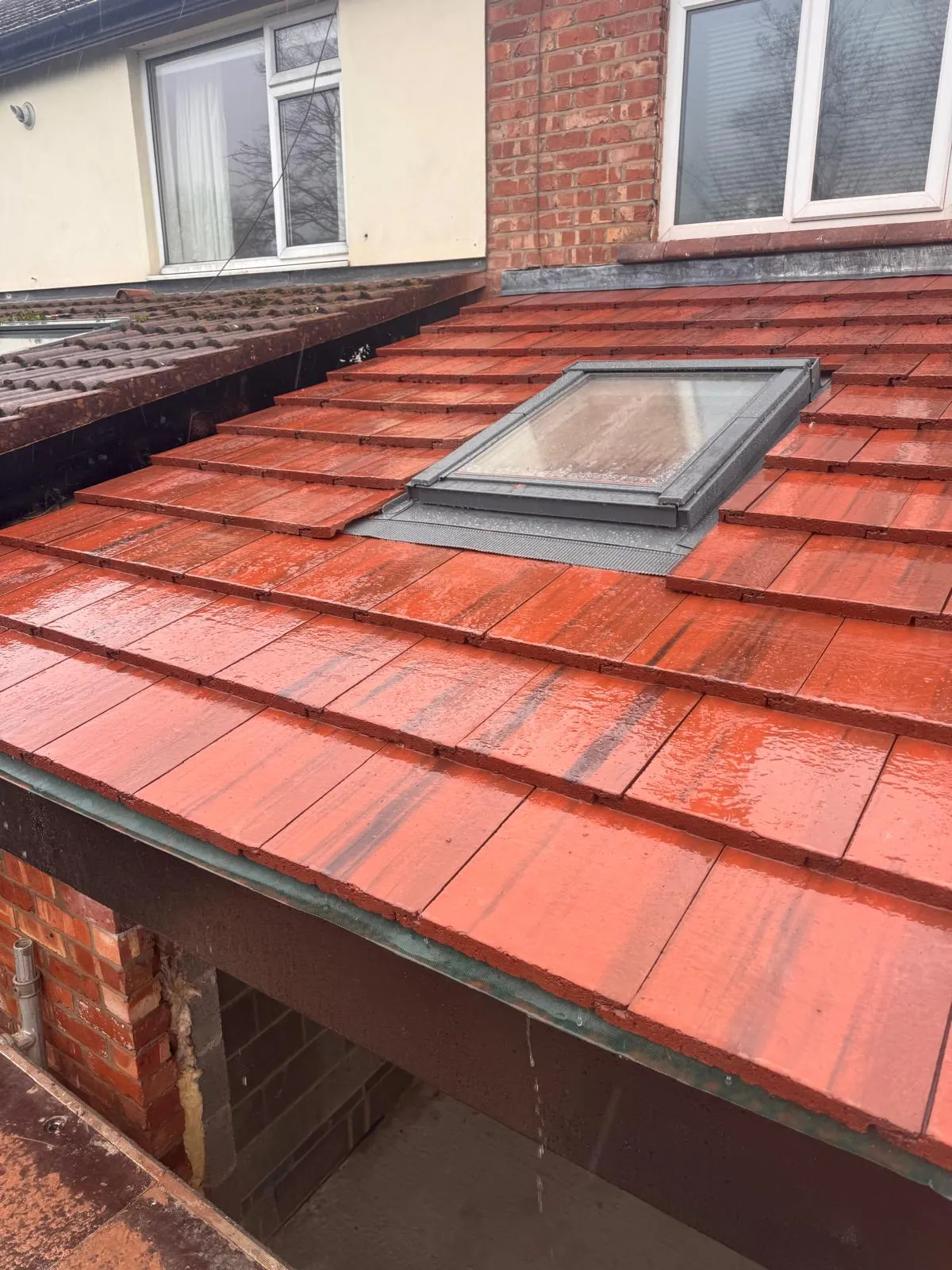 A close-up of a new residential roof with red tiles and a Velux window, wet from rain.
