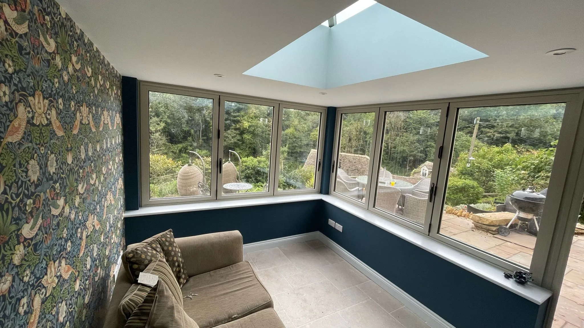 Bright orangery extension interior with sofa, patterned wallpaper, large windows, and skylight, built by DP Carpentry & Building LTD in Gloucestershire.