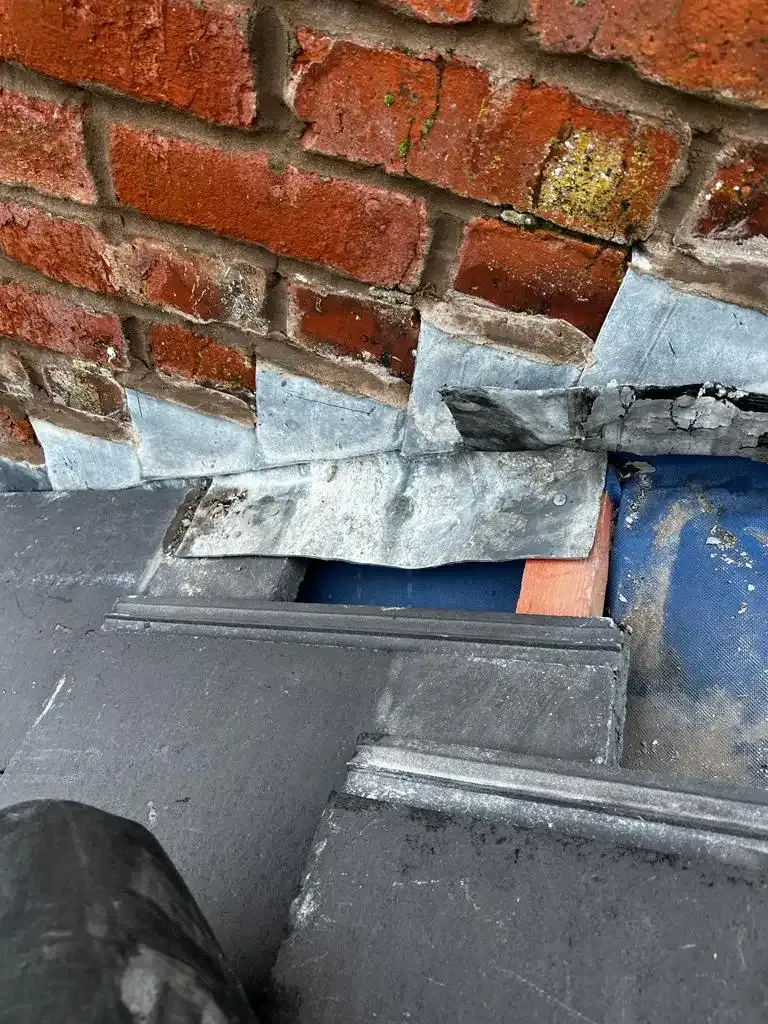 Damaged lead flashing on a tiled roof next to a brick wall, showing a gap.