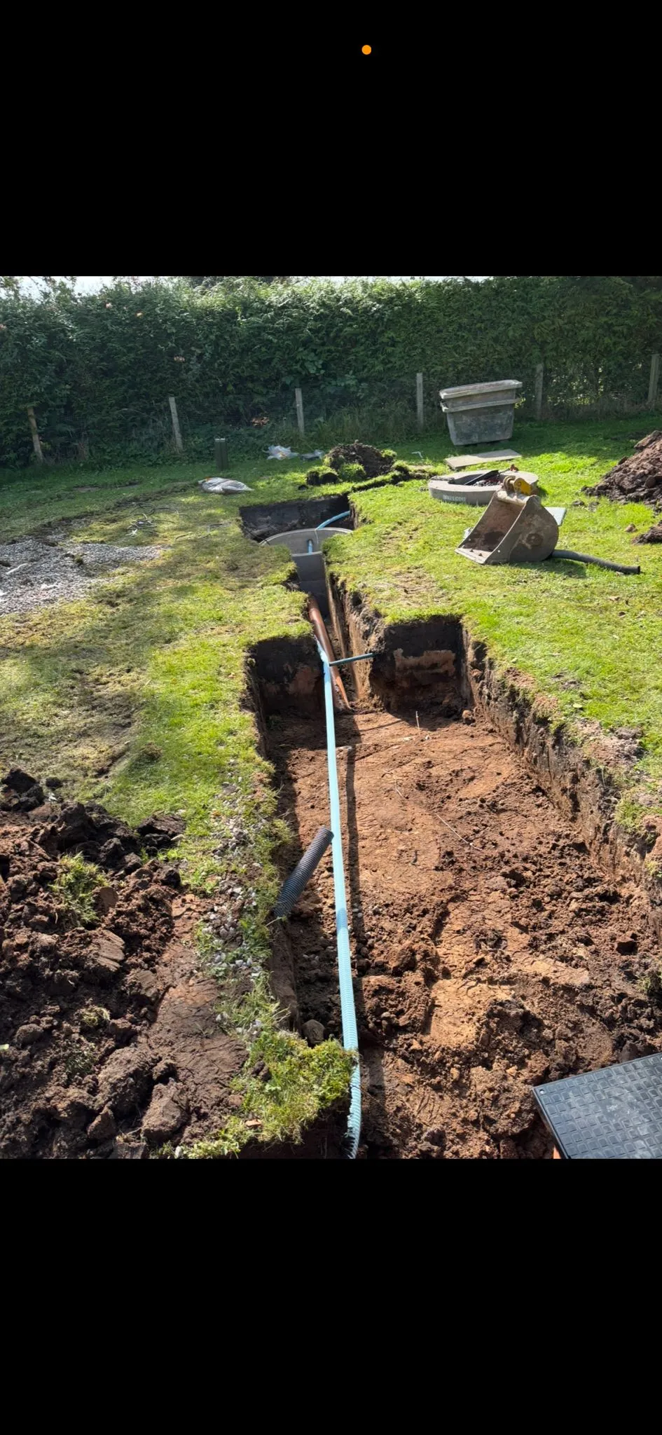 An open trench with blue and dark pipes laid, indicating drainage or utility installation in a grassy garden.