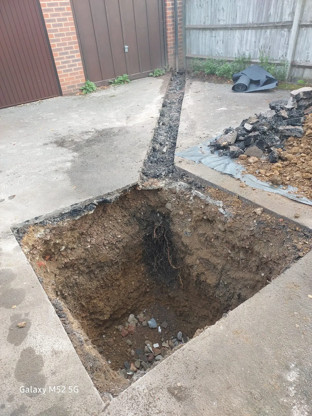 A construction site showing a deep square pit and a linear trench dug into concrete or tarmac, with piles of earth and debris nearby. Exposed tree roots are visible in the pit.
