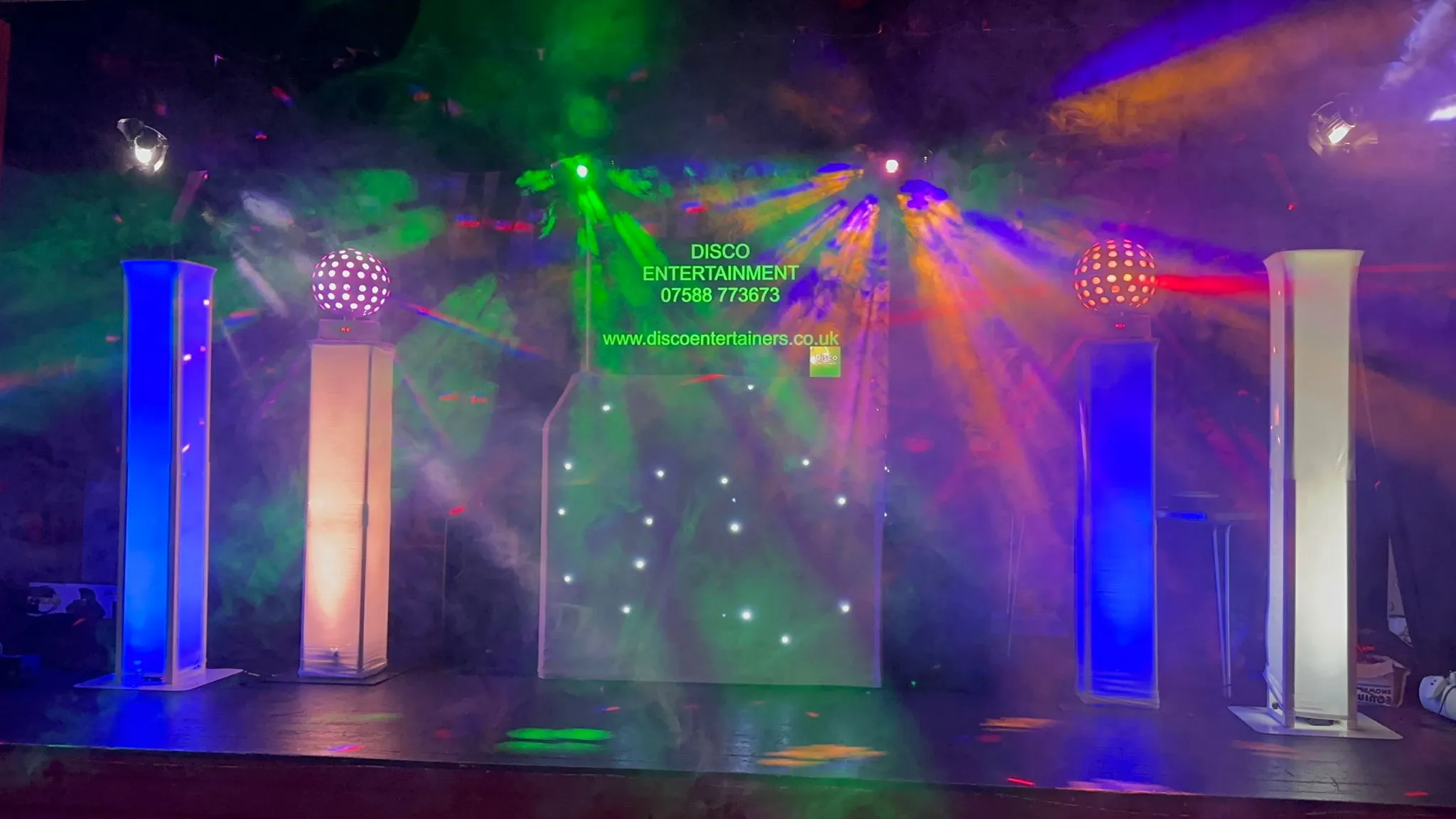 A vibrant DJ setup with colourful disco lights, two illuminated disco balls, and a central screen displaying company contact information.