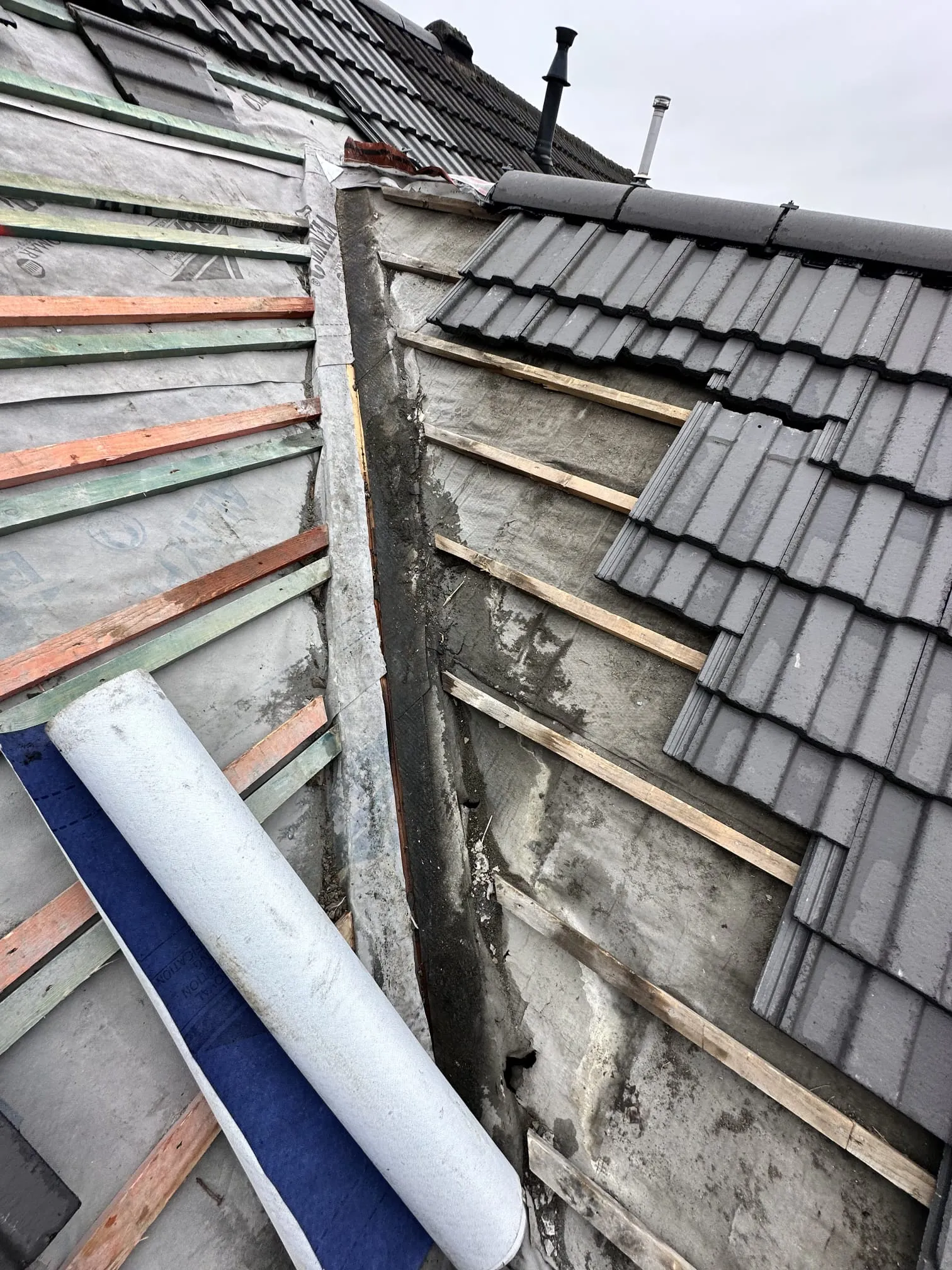 Close-up view of a roof being re-roofed, featuring new grey tiles, timber battens, waterproof membrane, and a roll of roofing material.