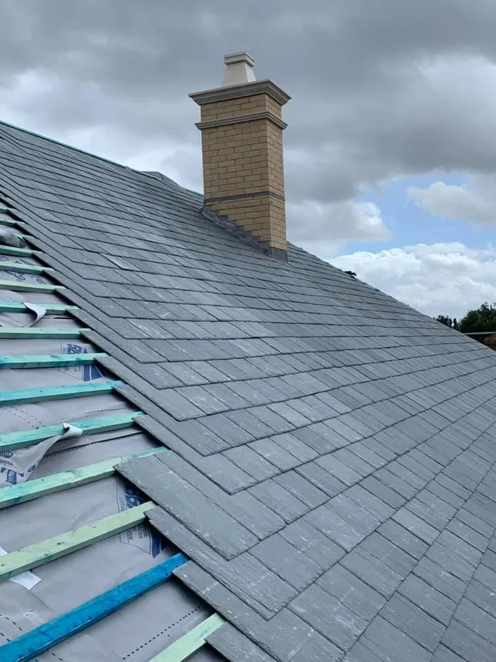 New grey roof tiles being installed on a pitched roof with visible underlay and battens next to a brick chimney.