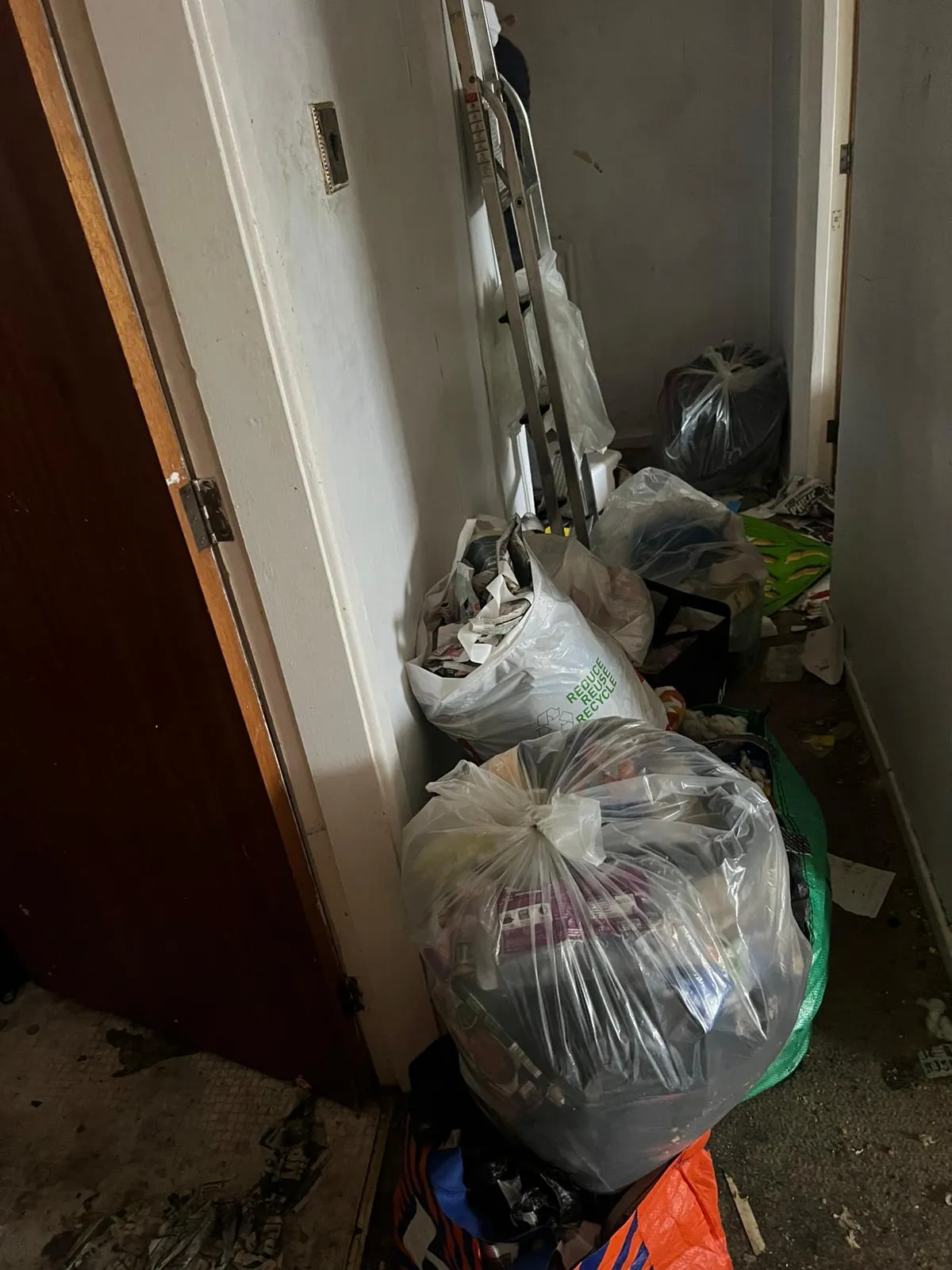 A cluttered residential hallway filled with bags of rubbish and a ladder leaning against the wall, indicating a hoarder clearance or general property cleanout.
