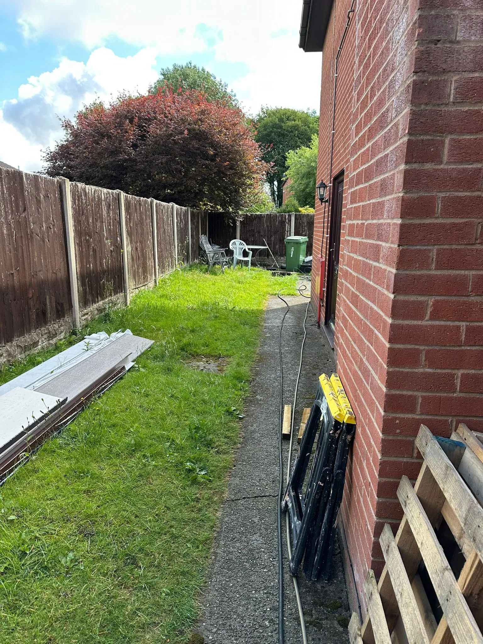 Side of a residential brick house with a path, grass, and a garden fence under a partly cloudy sky.
