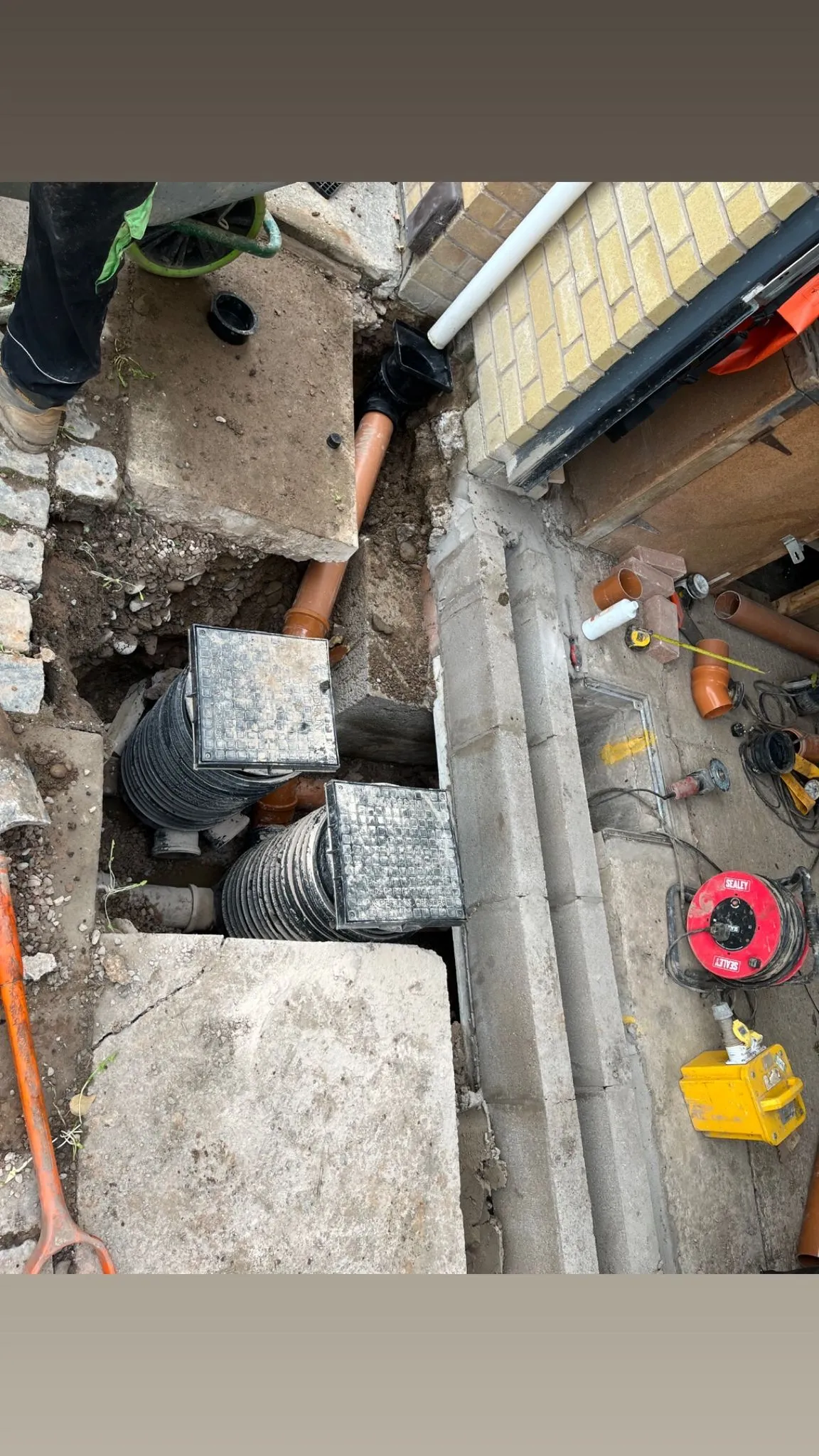 An overhead view of a drainage excavation project with orange pipes and manhole covers visible next to a brick building.