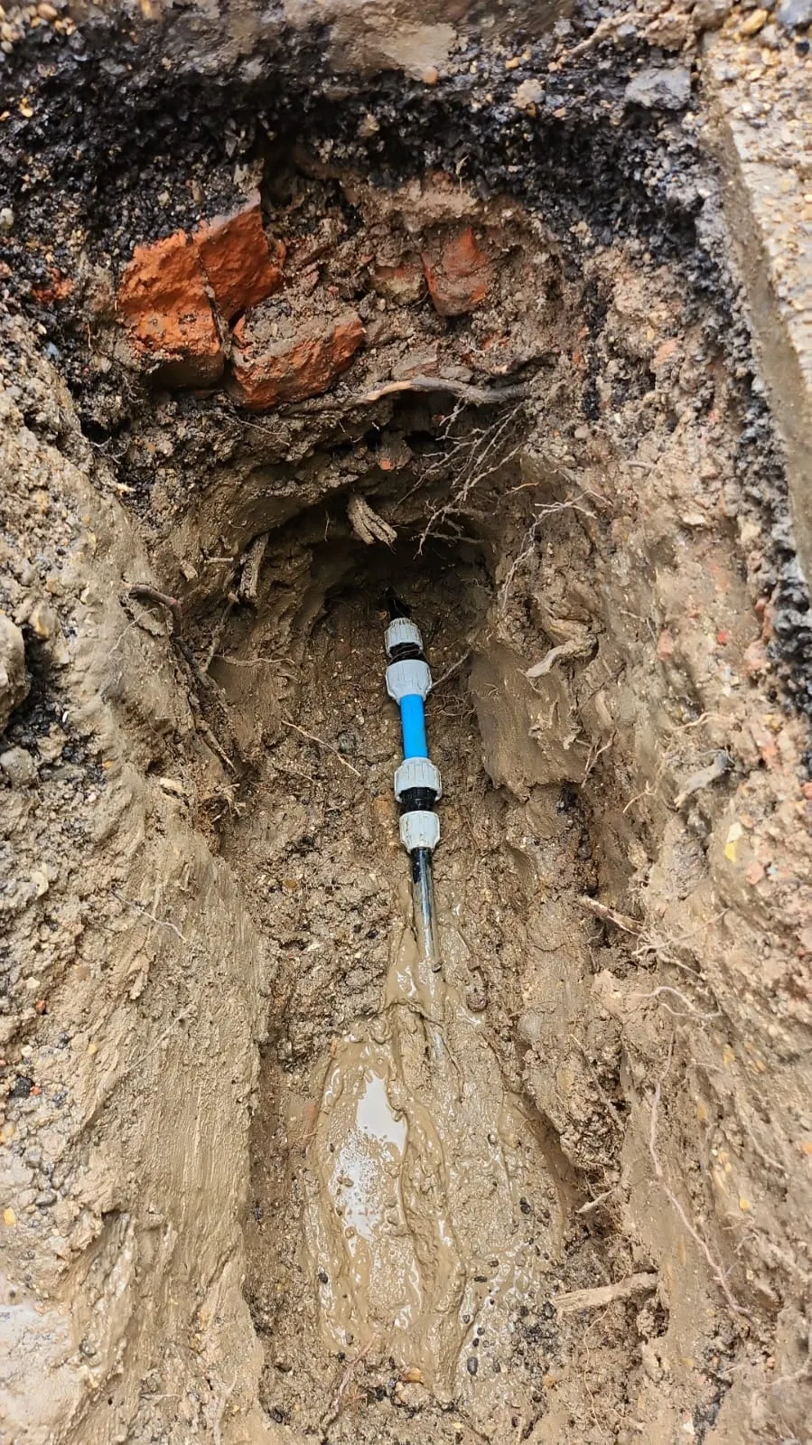 Close-up of an open trench dug into muddy ground, revealing a blue pipe segment with grey fittings, surrounded by soil, some bricks, and tree roots.