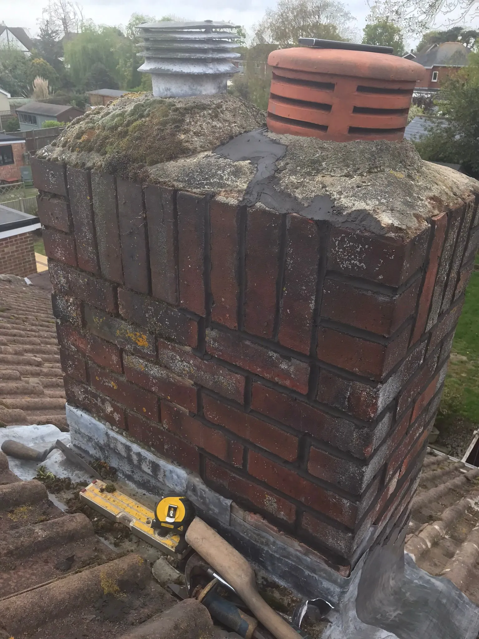 A brick chimney on a tiled roof showing moss, lead flashing, and tools for repair work