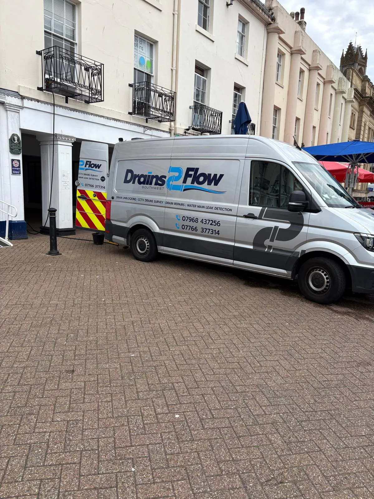 Drains2Flow branded service van with its rear door open, parked on a street in front of a building, with a hose extended towards the building, indicating active drainage work.