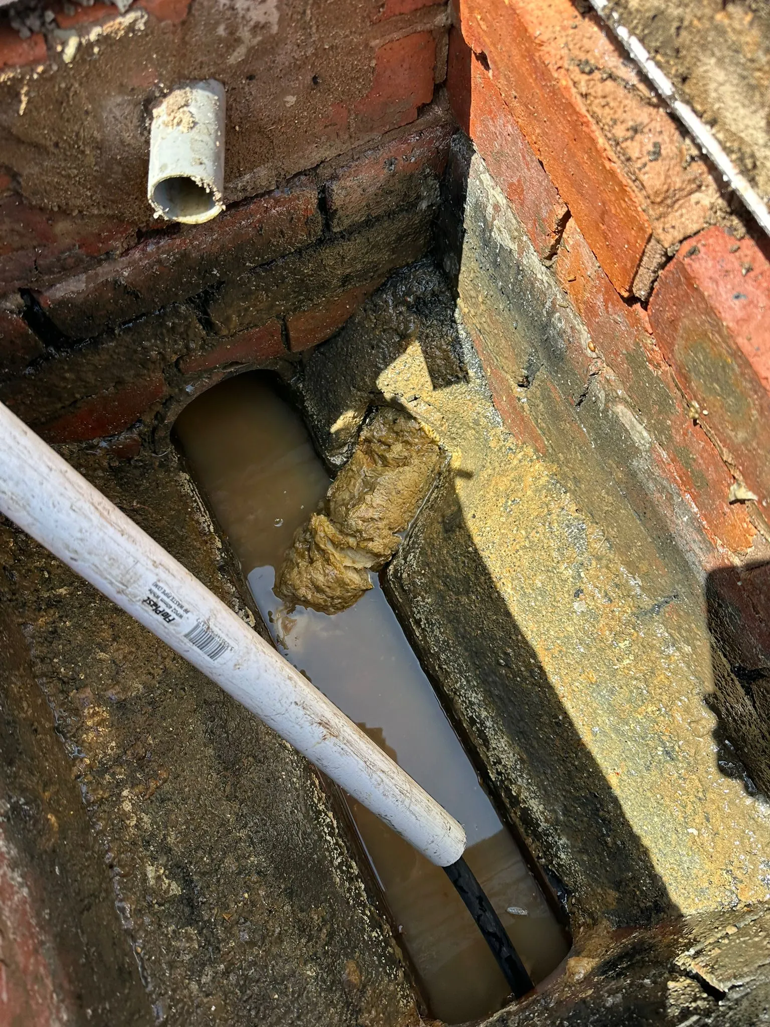 A close-up view of a severely blocked outdoor drain or manhole with dirty standing water and a large mass of fat or debris causing an obstruction.