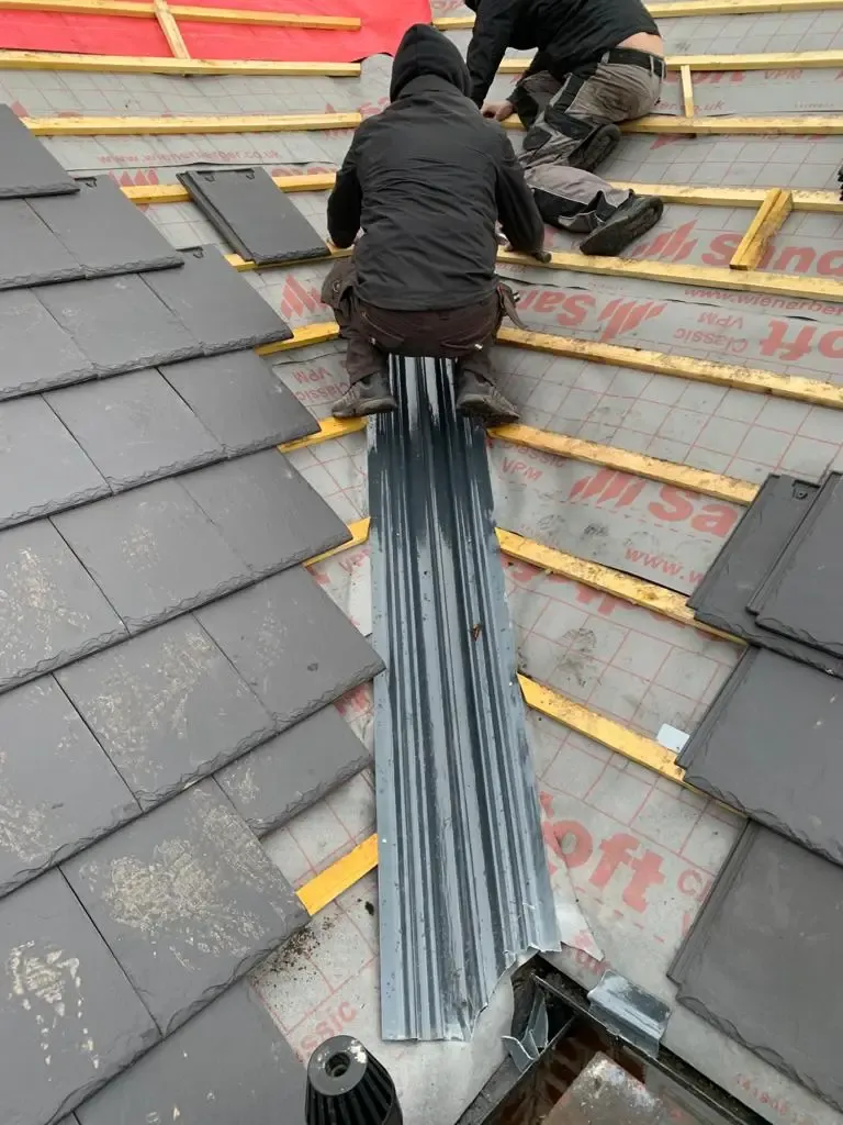 Two roofers installing slate or tile roofing with lead flashing on a residential property.