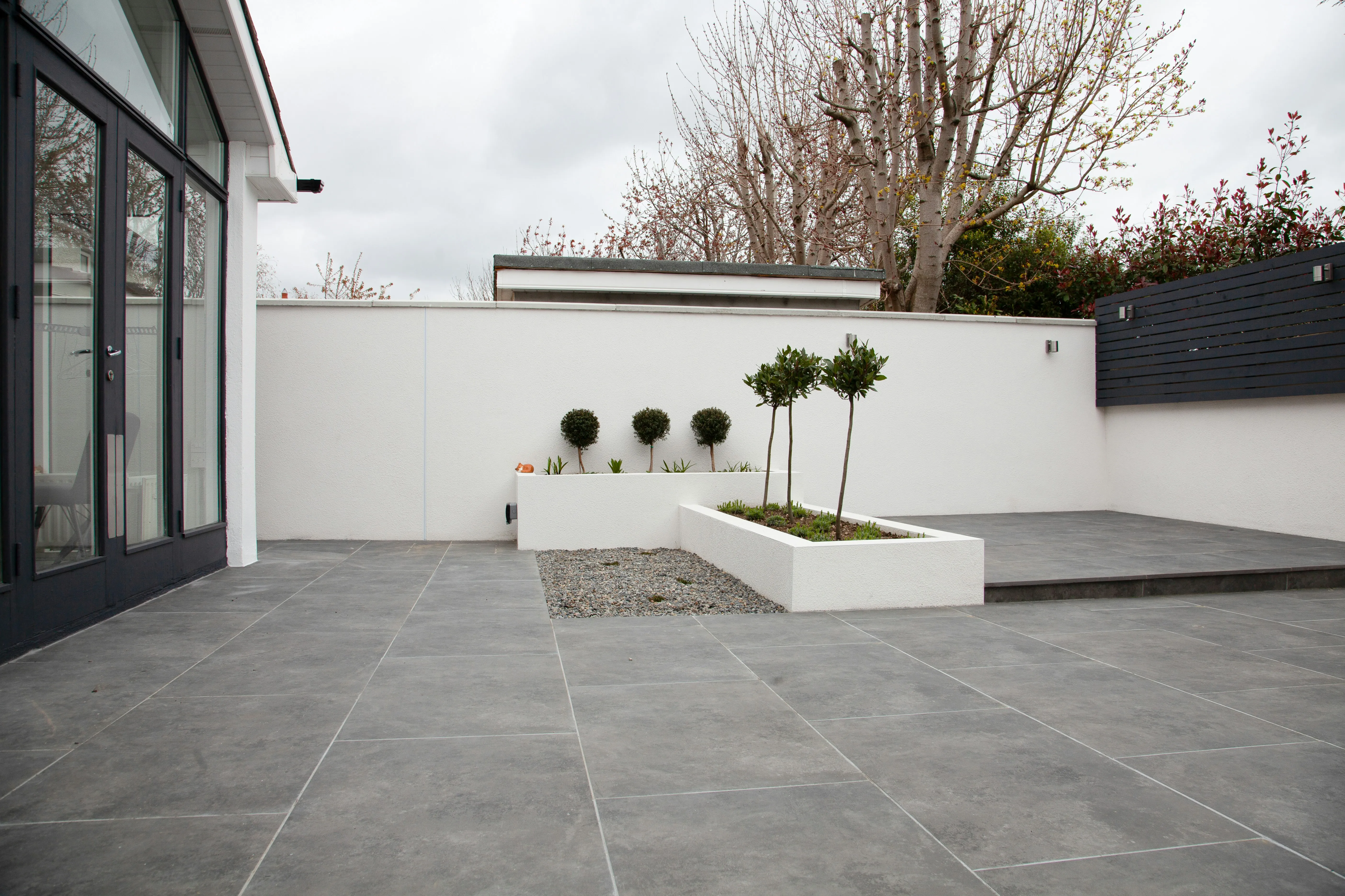 A modern grey patio with large format paving slabs, white rendered walls, and raised planters in a garden.