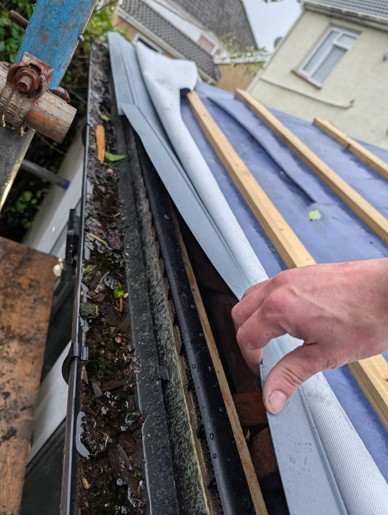 Close-up of a dirty, blocked gutter and a roofer's hand during a gutter and roofline replacement project.