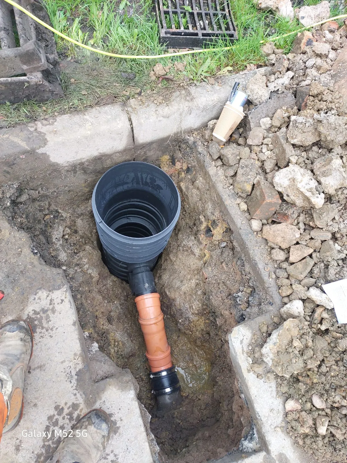 An excavated trench showing new orange and grey drainage pipes connected to a grey inspection chamber base during installation.