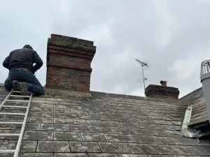 Roofer on a ladder repairing a brick chimney on a tiled roof.