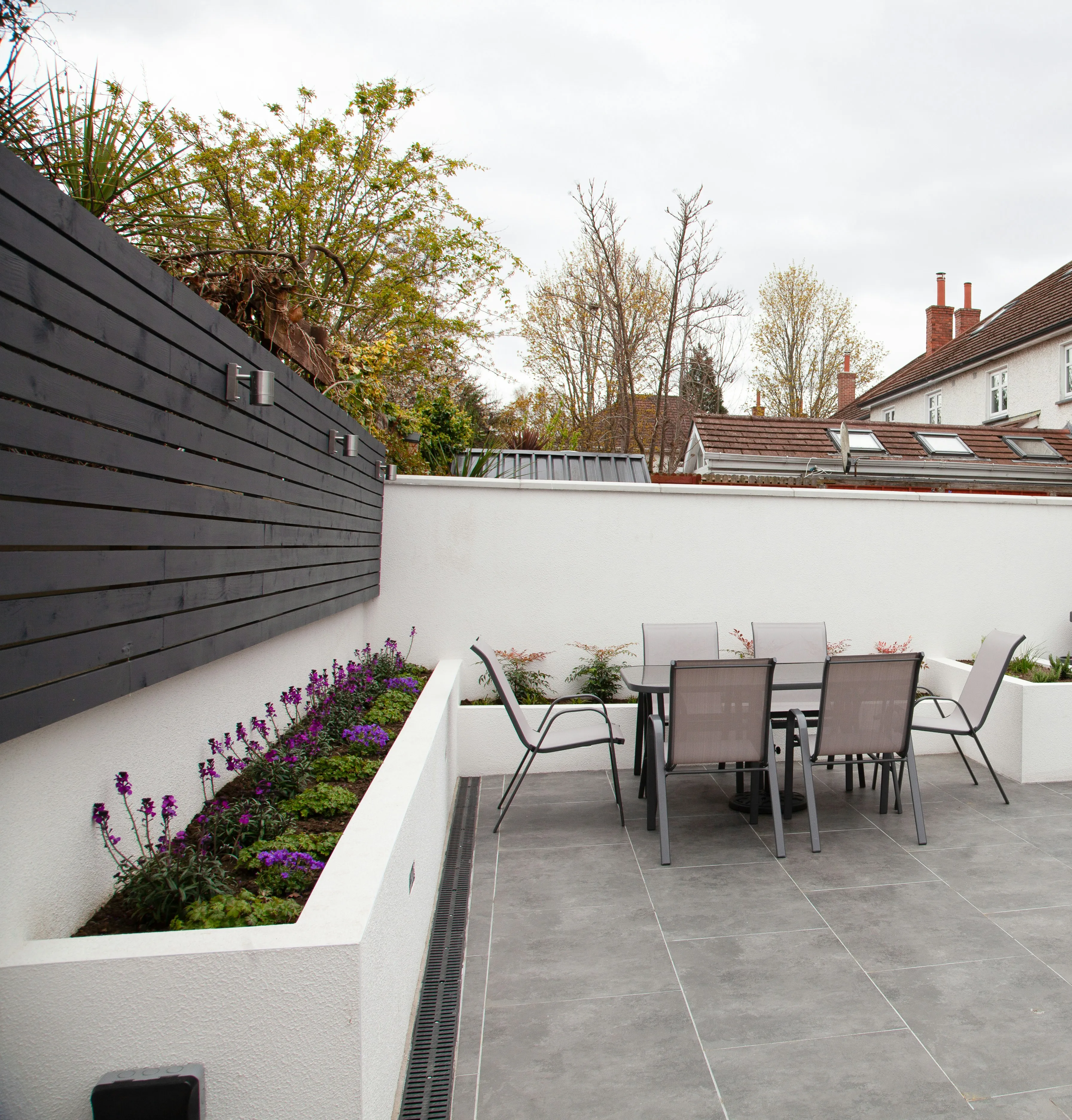 Modern grey paved patio with white raised walls, dark grey fence, garden furniture, and planted borders.