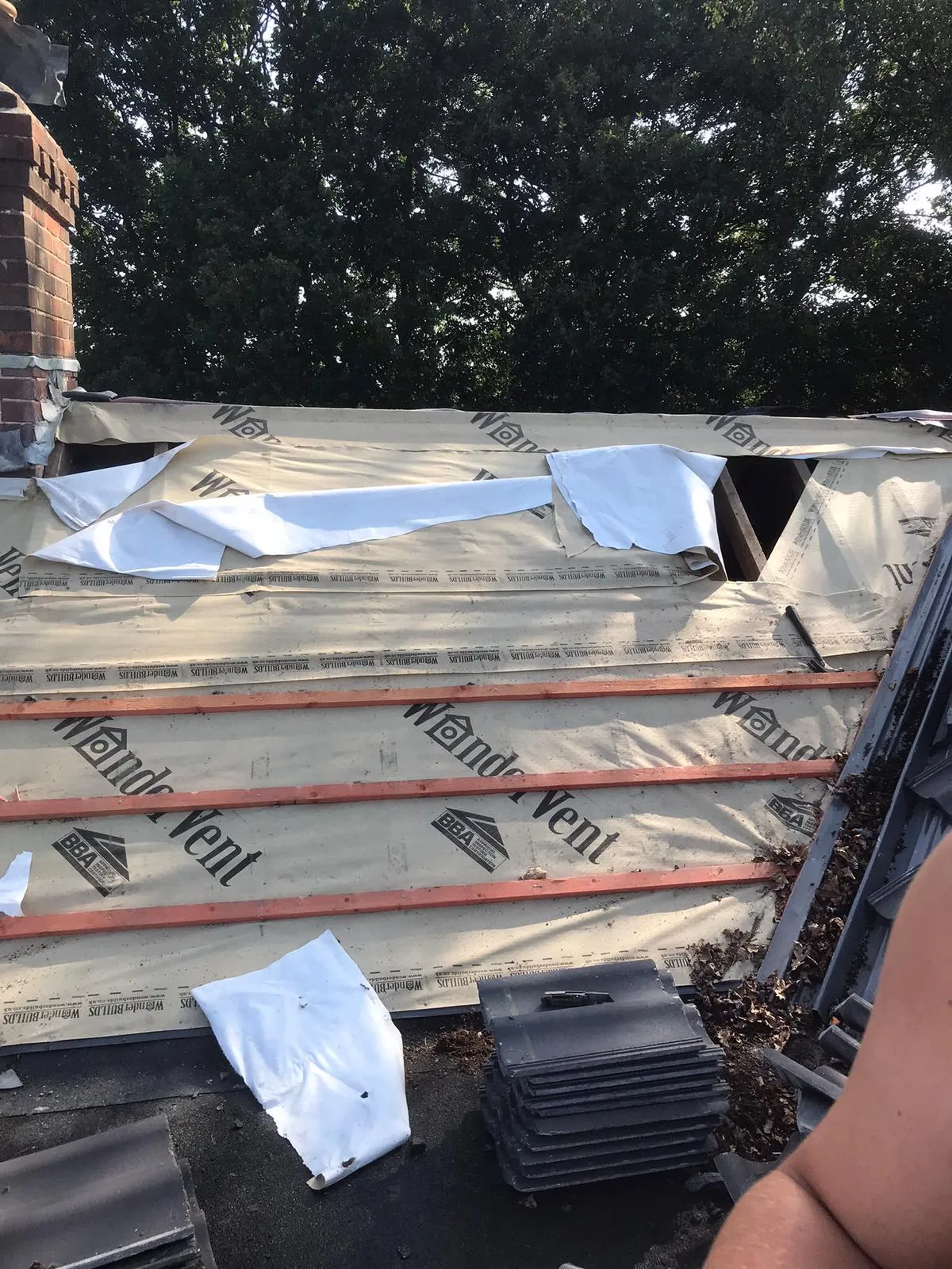 Close-up view of a pitched roof being re-roofed, featuring a cream-coloured breathable membrane with 'Wandervent' branding, horizontal wooden battens, and a pile of dark grey roof tiles ready for installation. A brick chimney is visible on the left, with green trees in the background under clear skies.