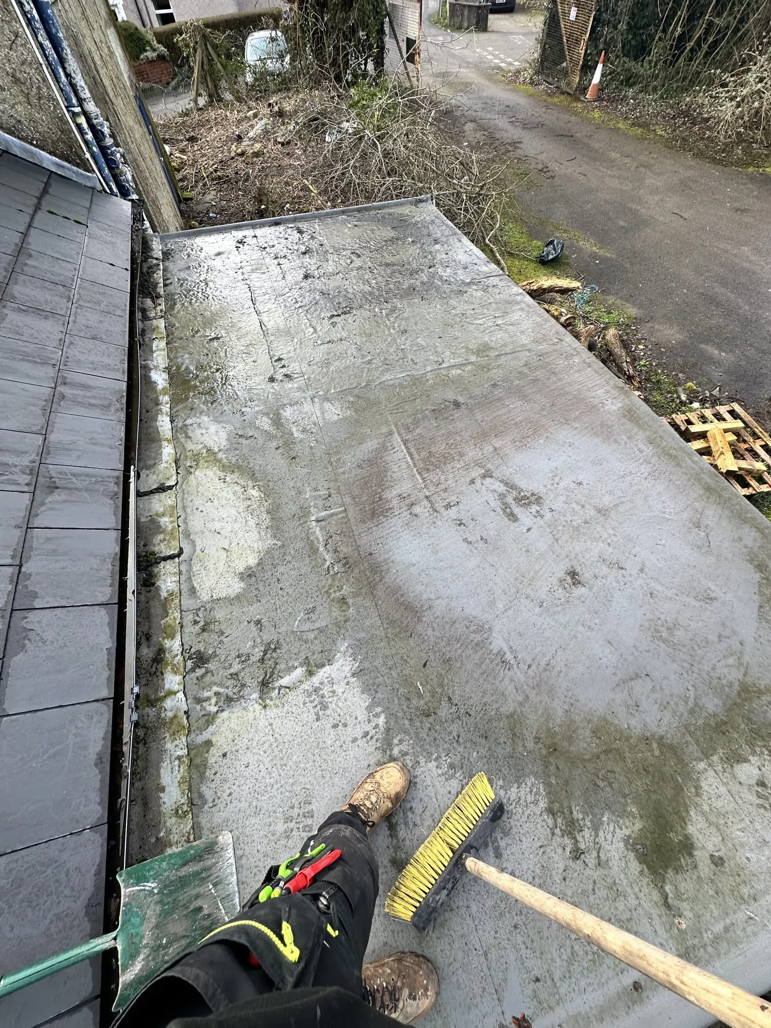 A dirty flat roof next to a pitched roof, with a roofer's boots and a broom, indicating roof cleaning work.