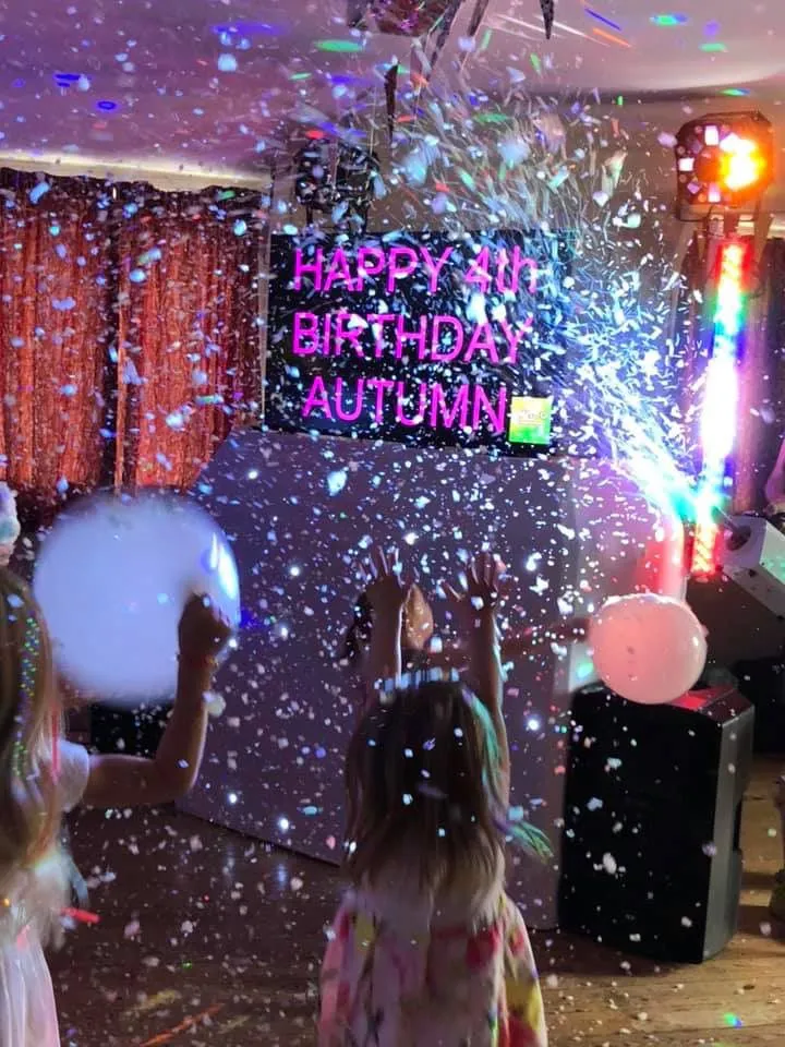 Two young children with their arms raised, delightedly playing in a shower of white confetti or 'snow' under colourful disco lights. A bright pink neon sign reads 'HAPPY 4TH BIRTHDAY AUTUMN'.
