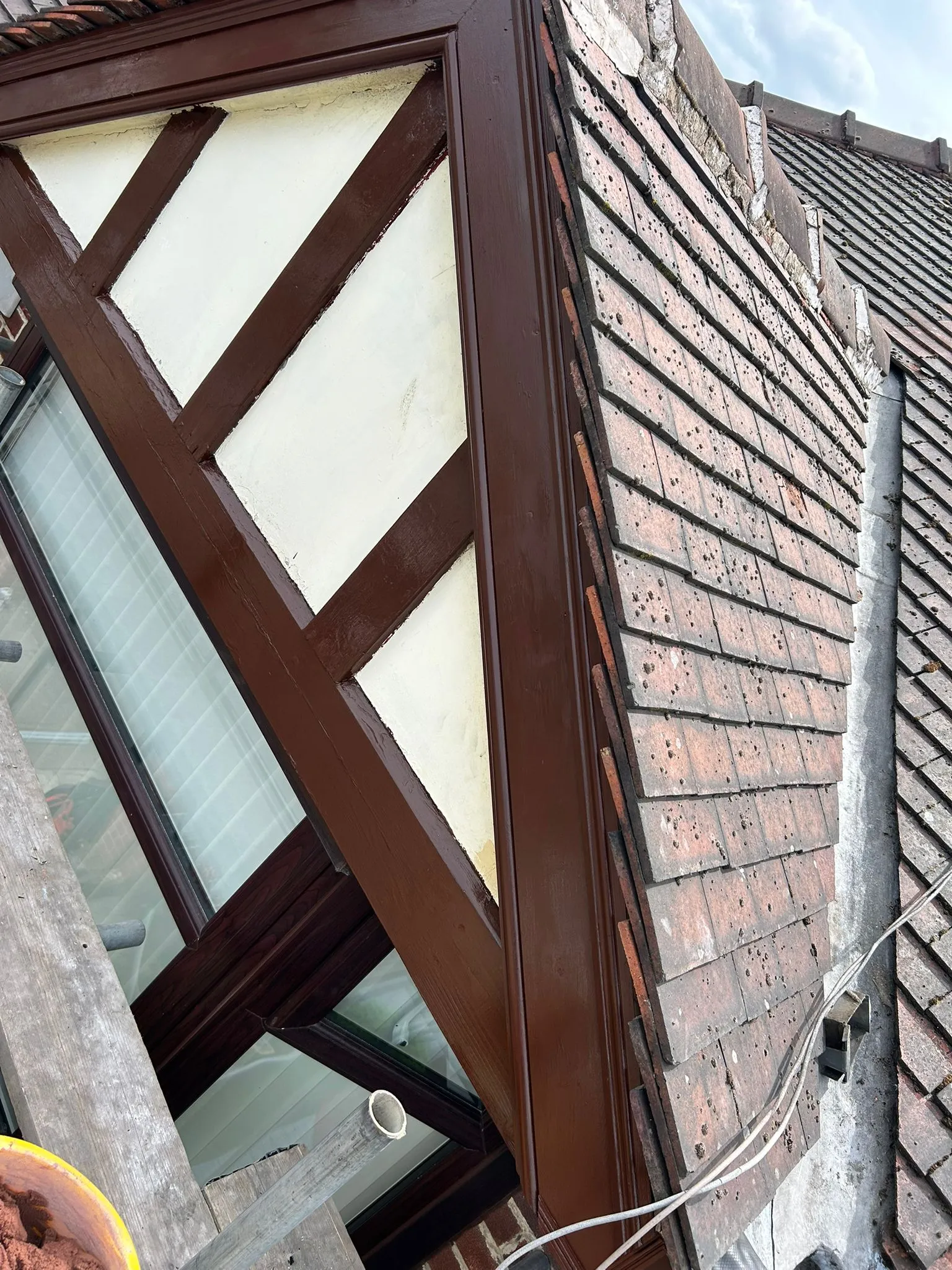 A close-up of a house roof showing old, weathered red tiles next to a brown-framed dormer window, suggesting roof repair or maintenance.