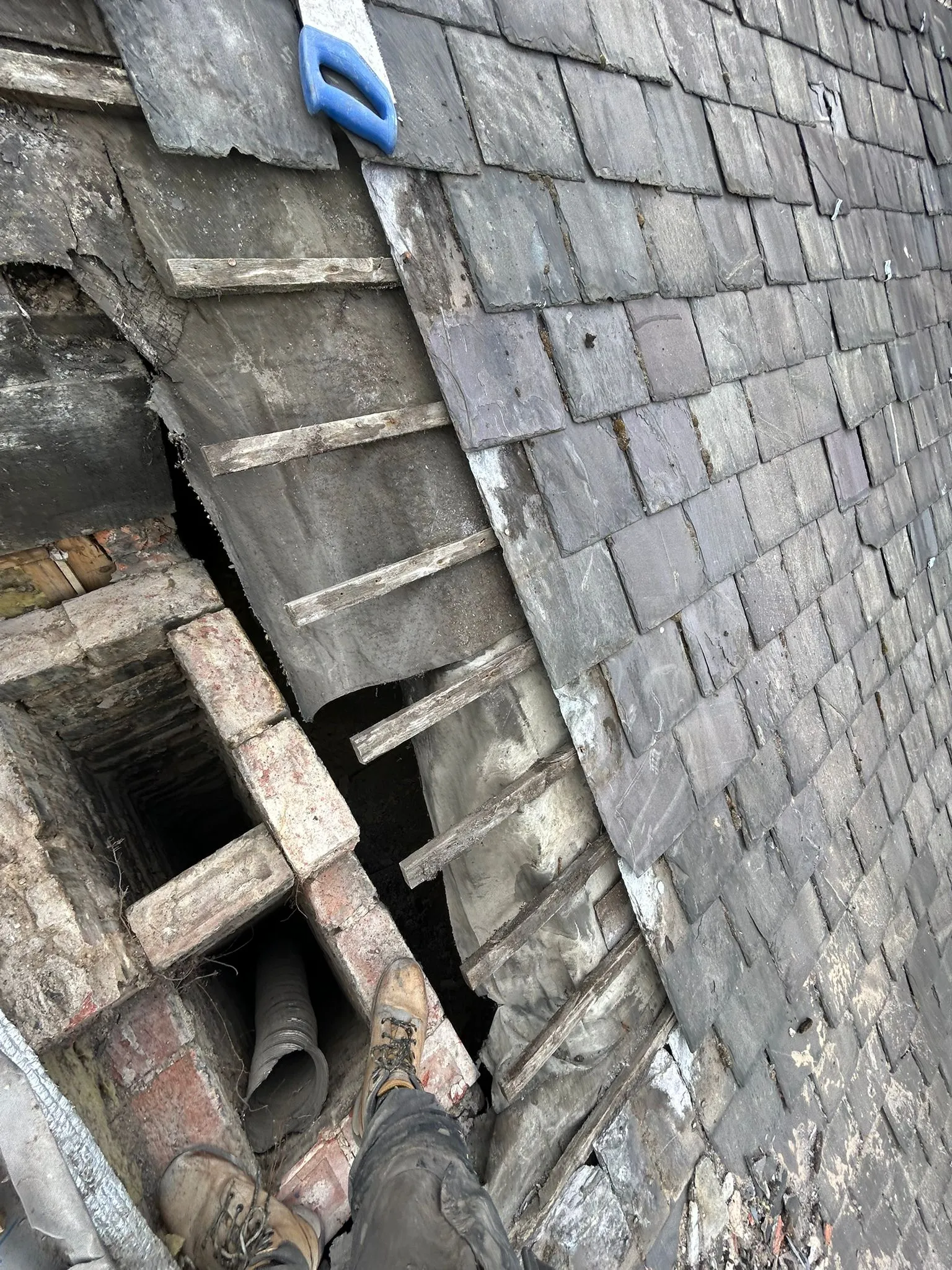 Damaged slate roof undergoing structural repair, showing exposed timber, chimney, and a roofer's boots.