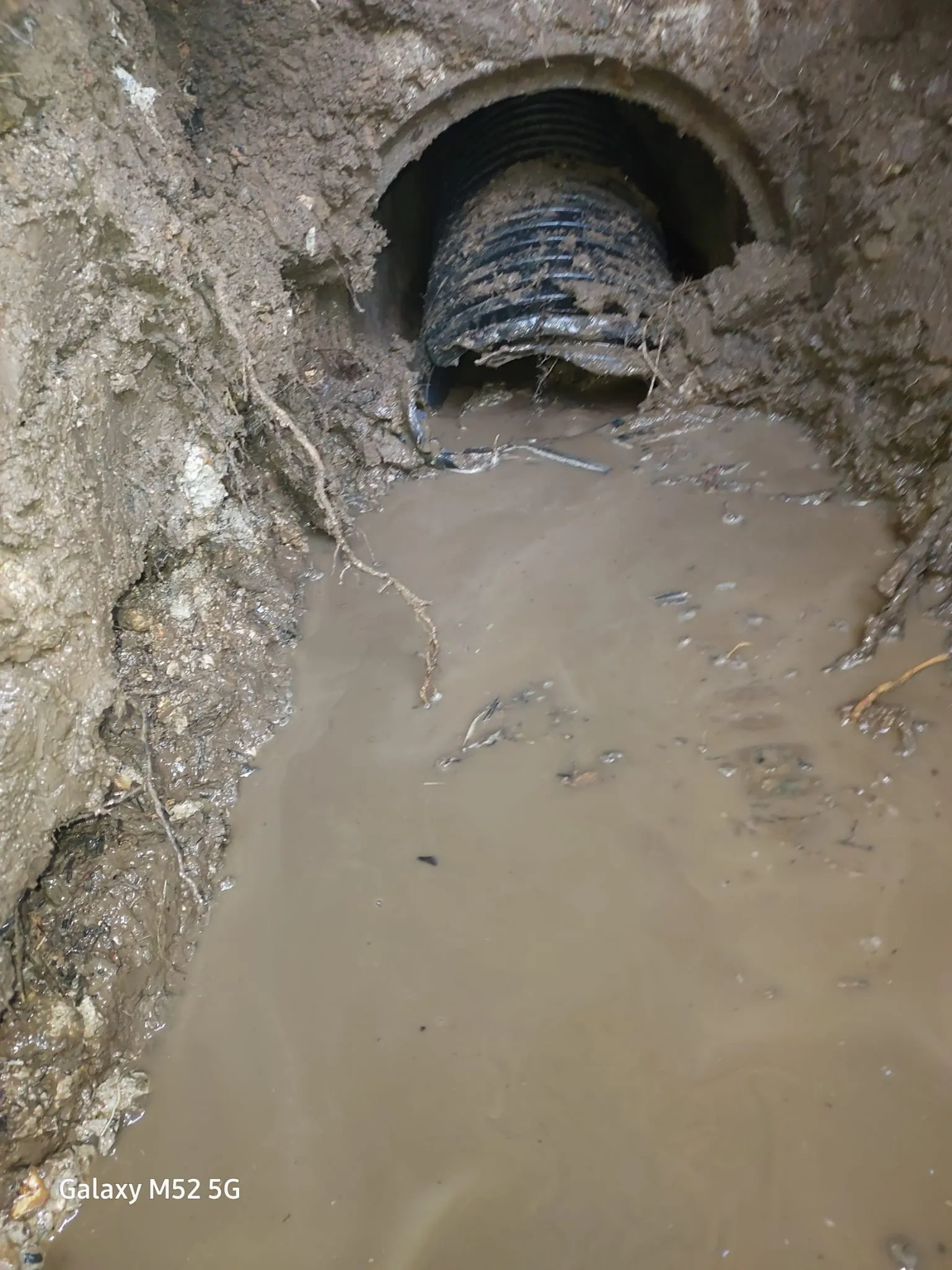 A blocked drain pipe surrounded by muddy water in an excavated trench, showing a drainage issue.
