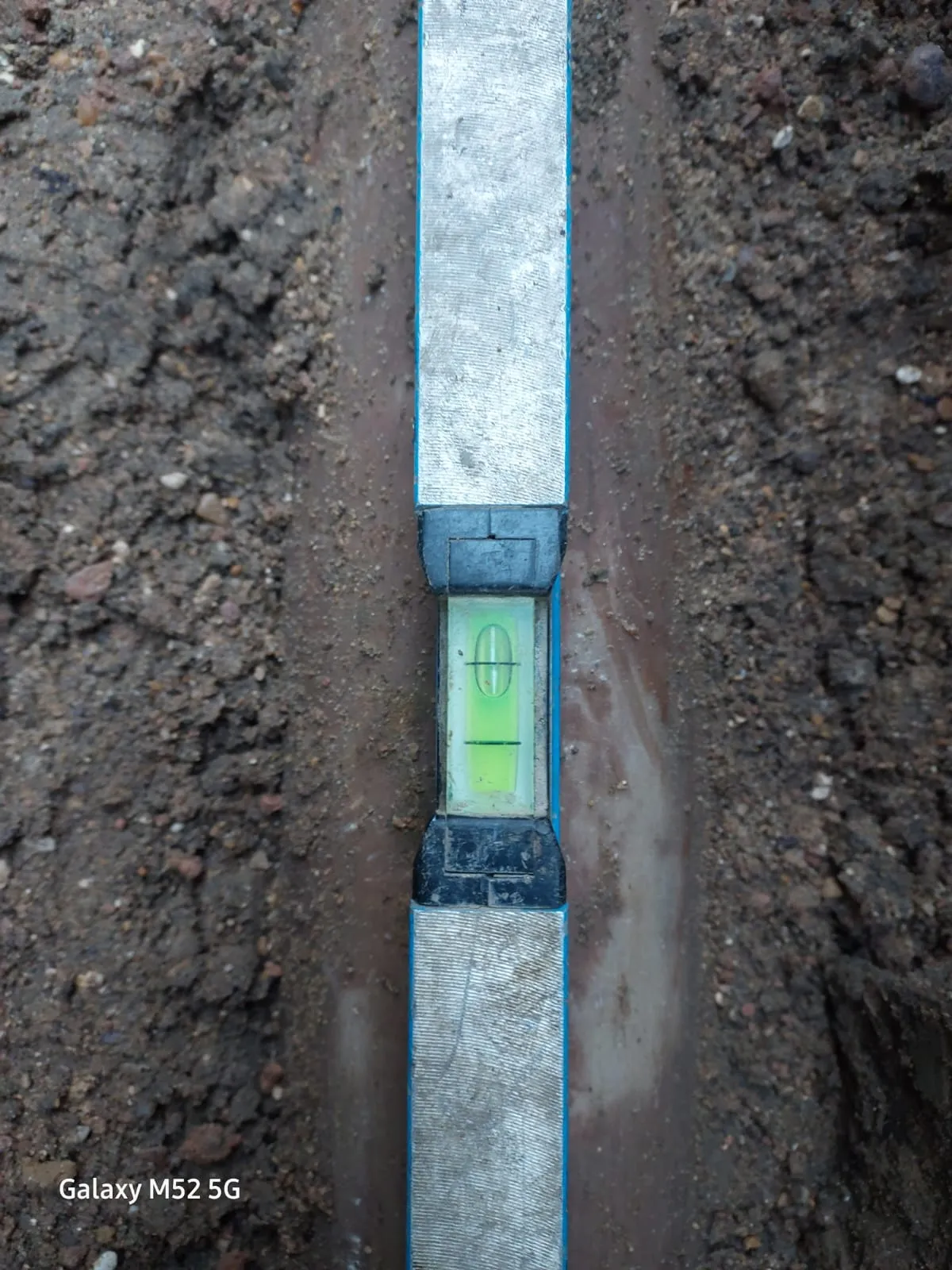 Close-up of a spirit level placed in a muddy trench, surrounded by soil, ensuring the correct angle for drainage pipework installation.