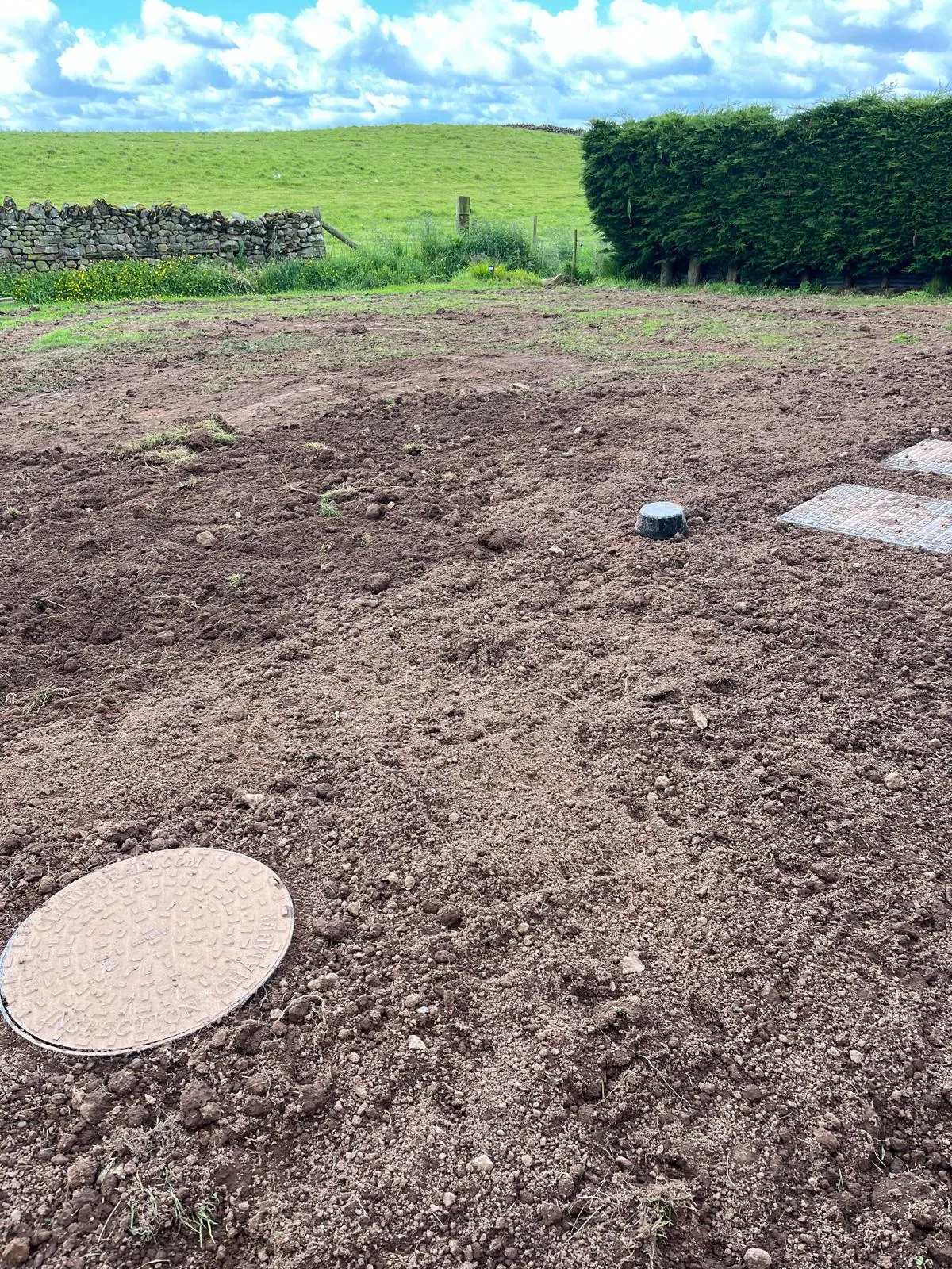 Recently excavated ground for drainage or septic tank installation, featuring a round inspection chamber manhole cover and square access points.