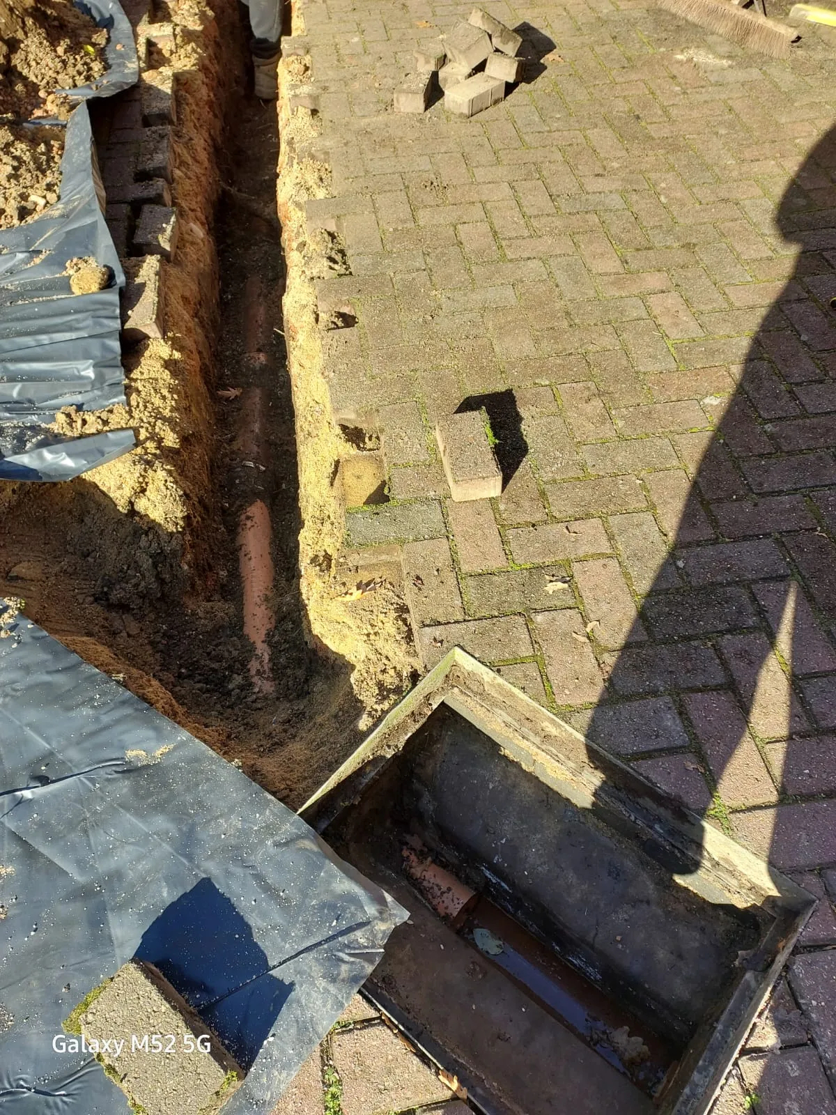An excavated trench showing an orange drain pipe with an open manhole cover nearby, set against block paving.