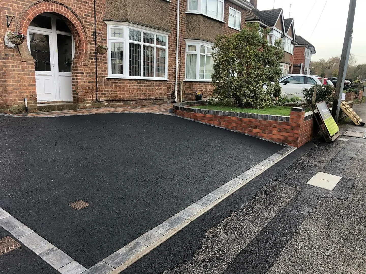 New tarmac driveway with grey block paving border in front of a red brick house and a small lawn with a low brick wall.