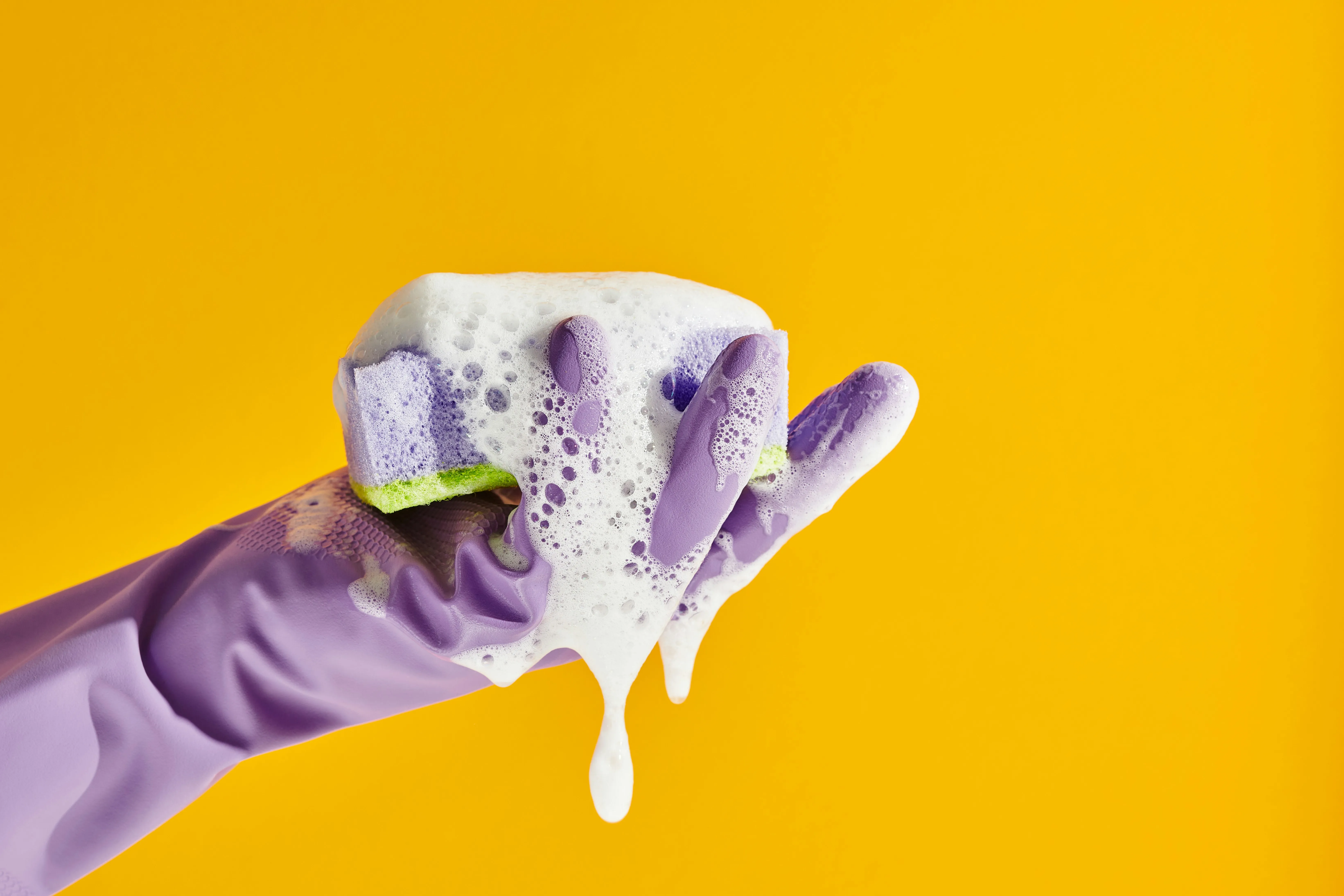 A hand in a purple rubber glove holding a sponge covered in white soap suds, on a bright yellow background.