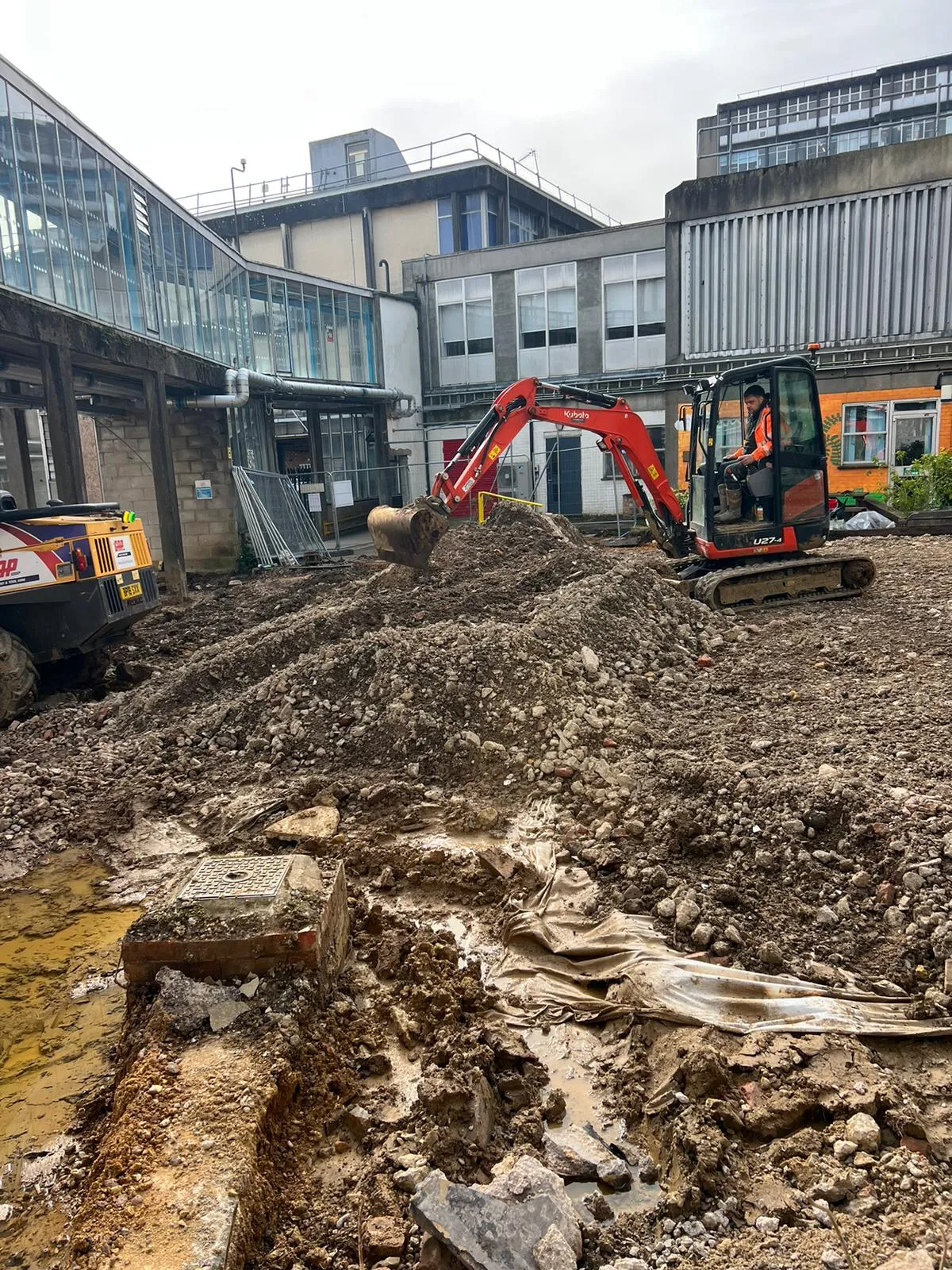 A red Kubota excavator digging a large pile of earth at a commercial construction site, with multi-story buildings in the background and an exposed drain access point.
