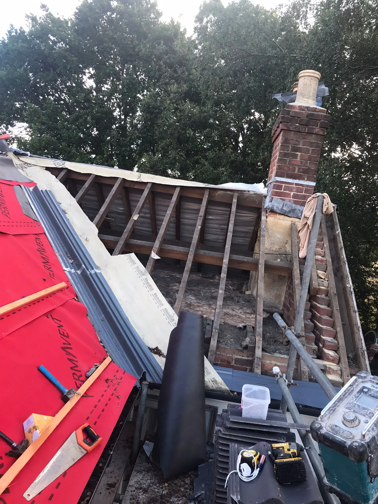 Roof installation or major repair in progress, with visible rafters, red roofing underlay, a brick chimney, and construction tools.