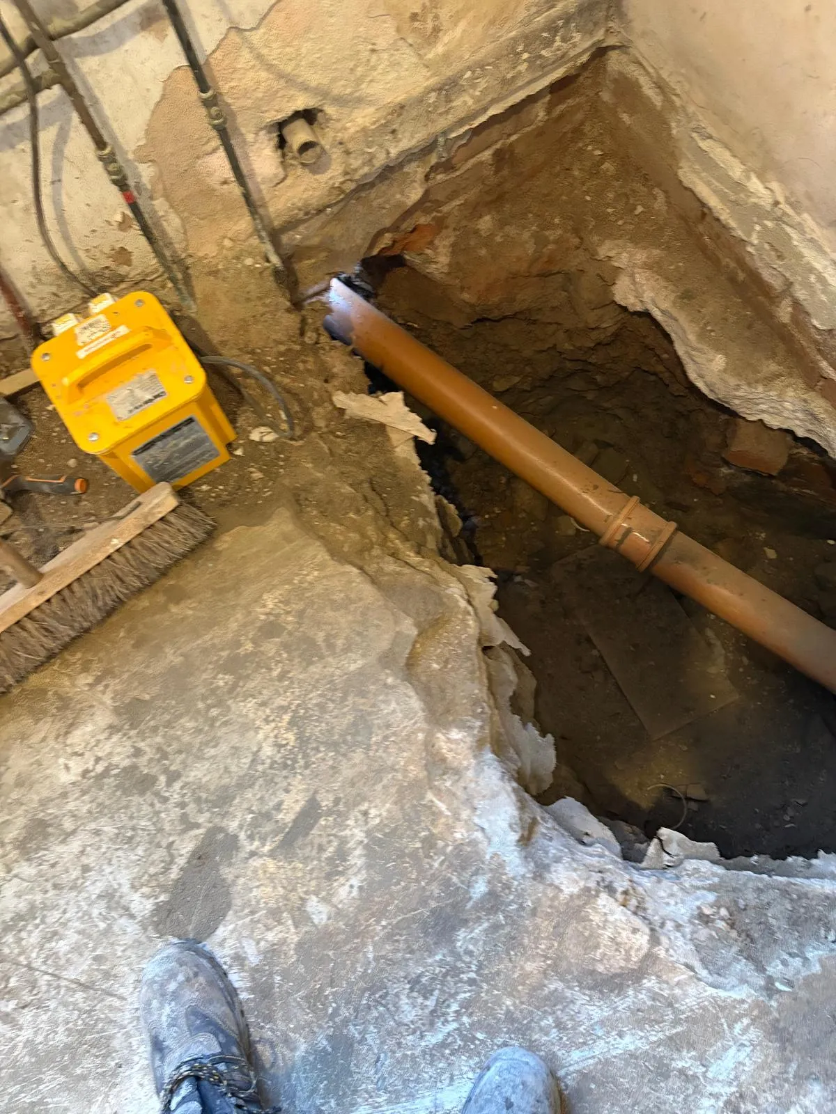 Excavated floor exposing a drainage pipe for repair or replacement, with a pipe locator tool on the ground.