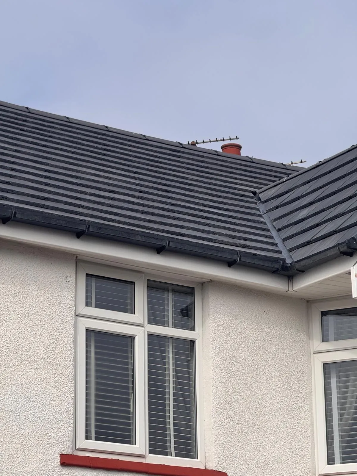 Close-up of a residential building's dark tiled roof, white uPVC windows, and textured white wall, with a chimney pot and aerial.