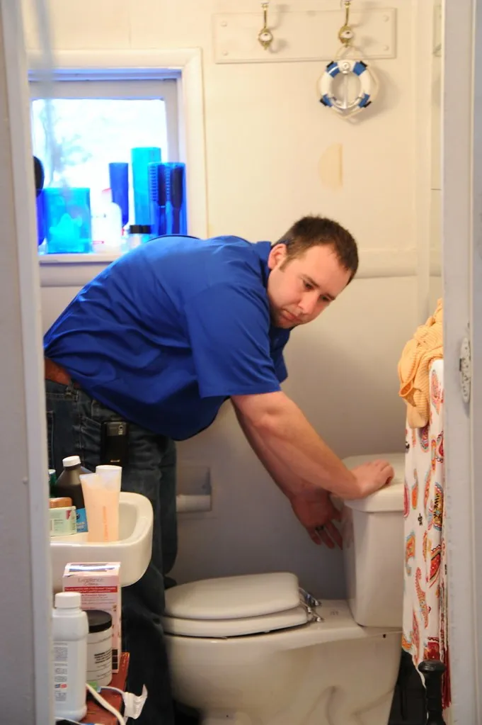 A male engineer in a blue shirt working on a toilet in a home bathroom, representing emergency toilet unblocking or repair.
