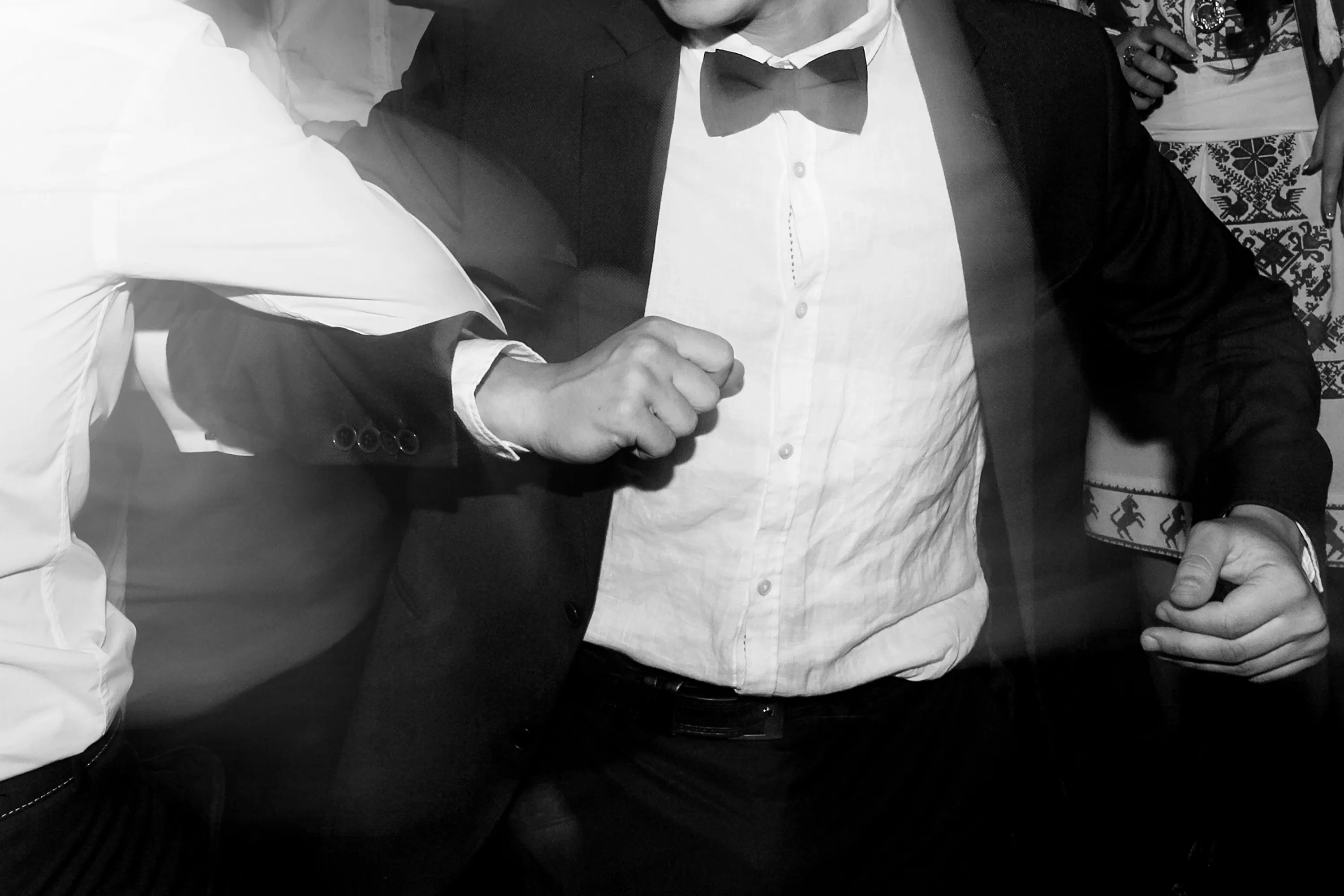 A black and white photo of two men in formal wear, one in a suit and bow tie, dancing enthusiastically at a lively party or wedding, showing joyful celebration on the dance floor.
