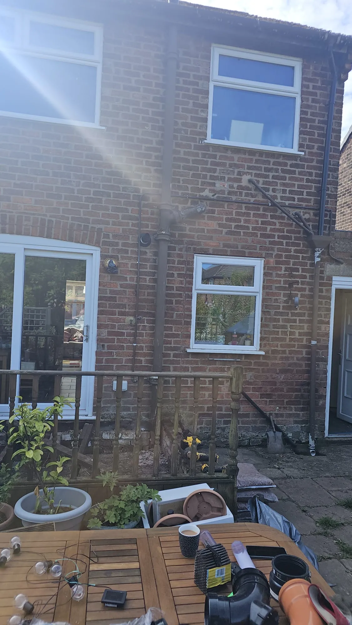 Rear of a brick house with external drainpipes and new plumbing parts on a patio table, indicating drainage work.