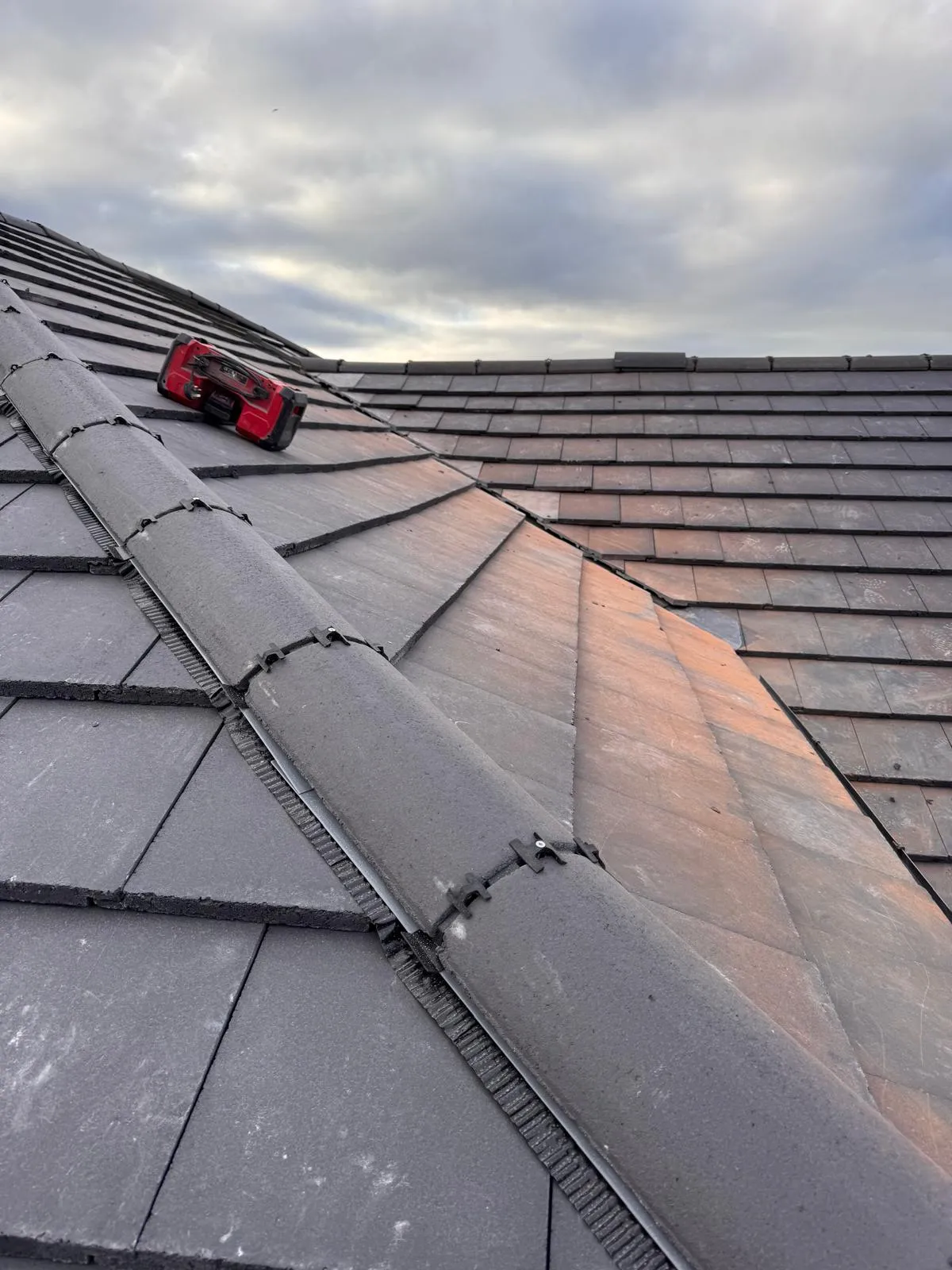 Dark grey tiled roof section with ridge tiles and a red power tool, illustrating professional roofing work.