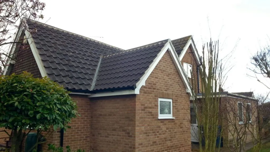 A residential house with a dark pitched tiled roof and brick walls. The roof has gabled sections and white roofline trim. Trees and bushes are visible around the property.
