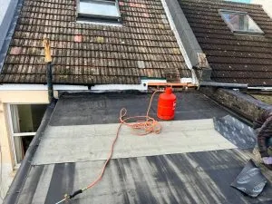 Roofers working on a flat roof, applying roofing felt with a gas torch, with a tiled pitched roof and Velux windows in the background.