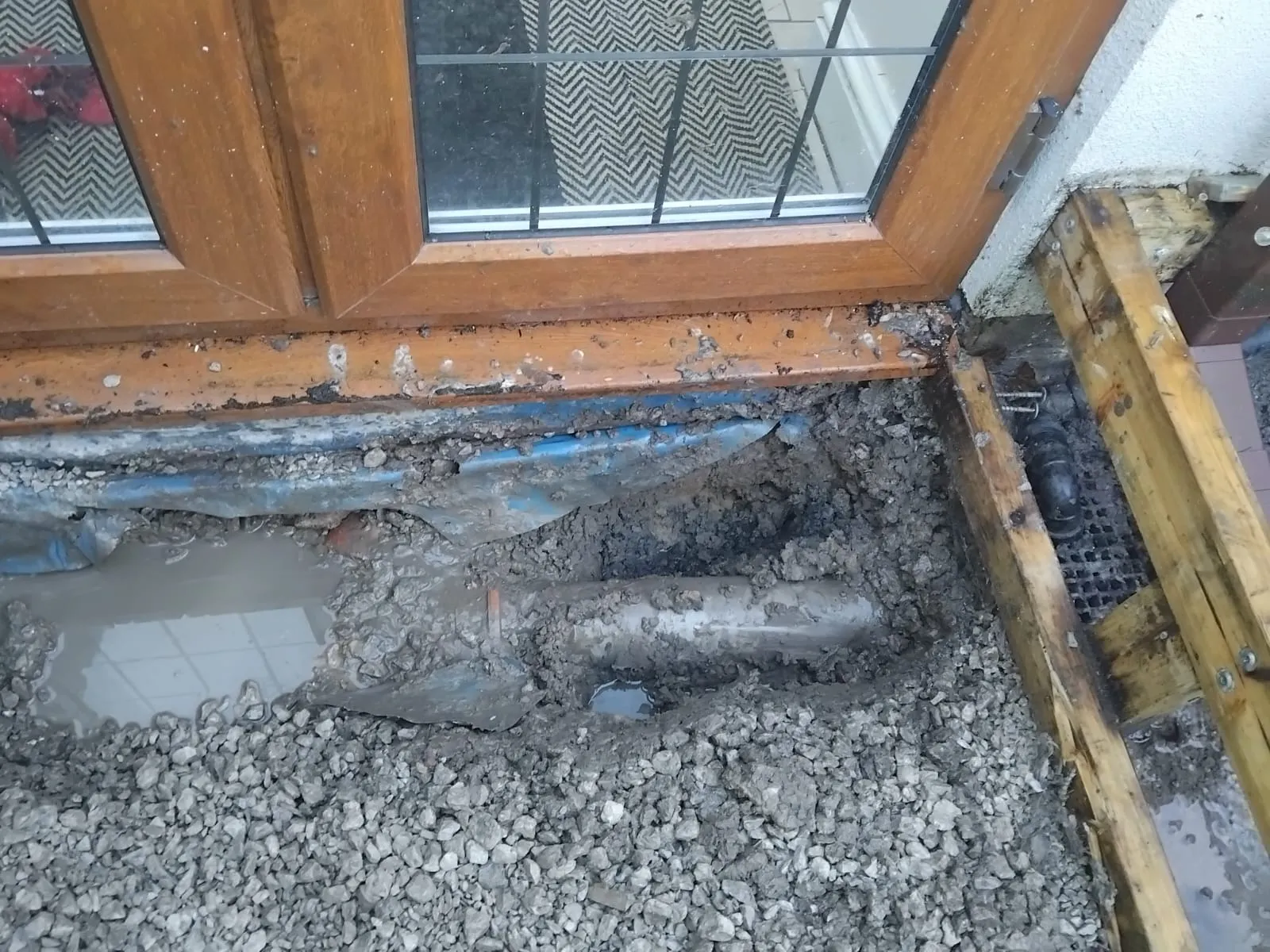 Image showing an excavated trench with exposed drainage pipes, gravel, and muddy water next to a building's entrance with a brown doorframe.