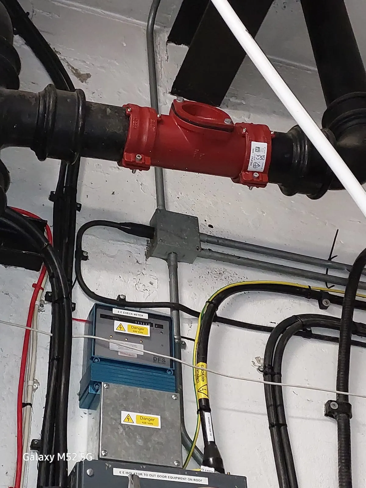 Close-up of a red non-return valve on a black pipe, with electrical conduits and meters in the background, likely in a commercial or industrial setting.