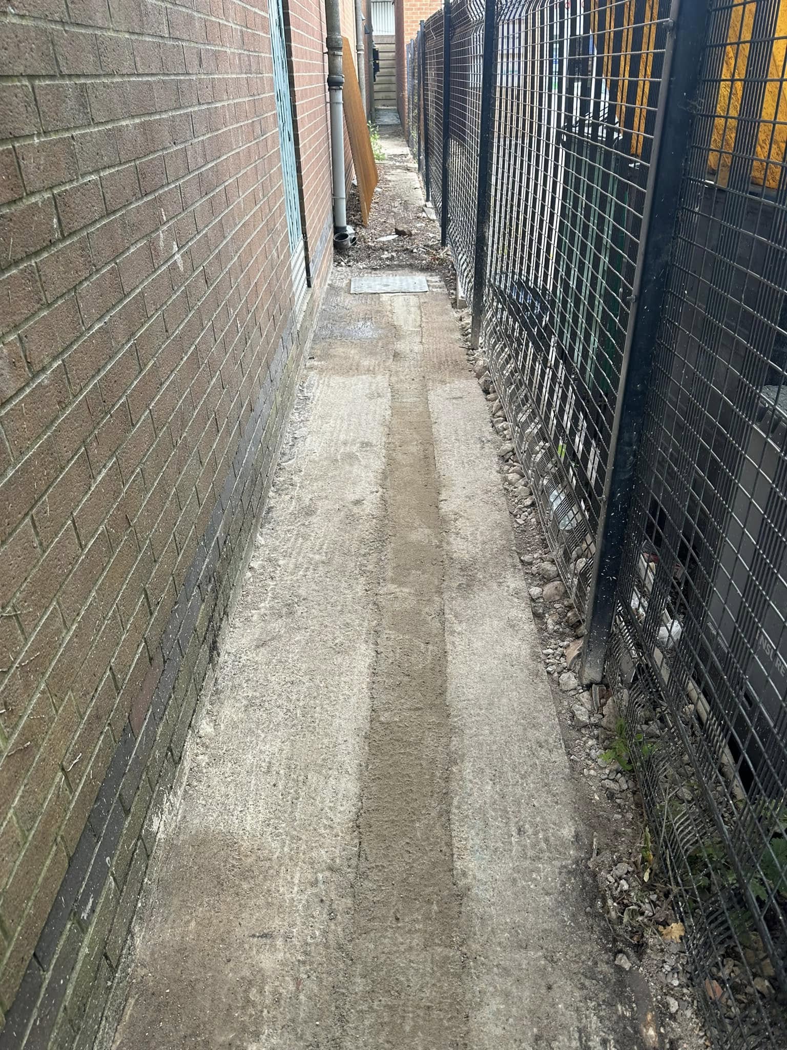 Narrow concrete path next to a brick building and metal fence, an area for drainage.
